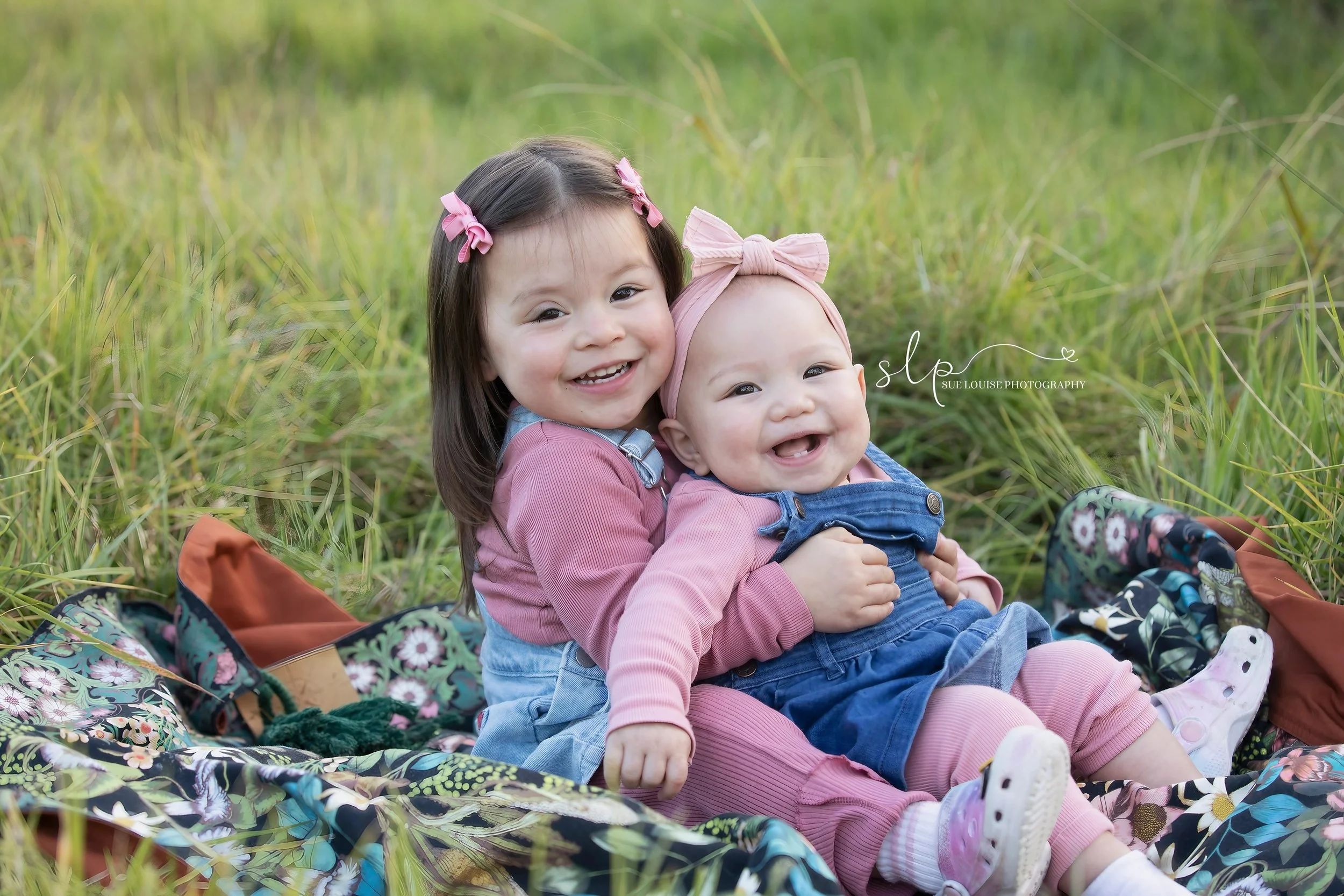 Two young girls sitting on a floral blanket in a grassy field, smiling at the camera. One girl has dark hair with pink bows, and the other has light hair with a pink headband, both wearing pink and denim clothing.