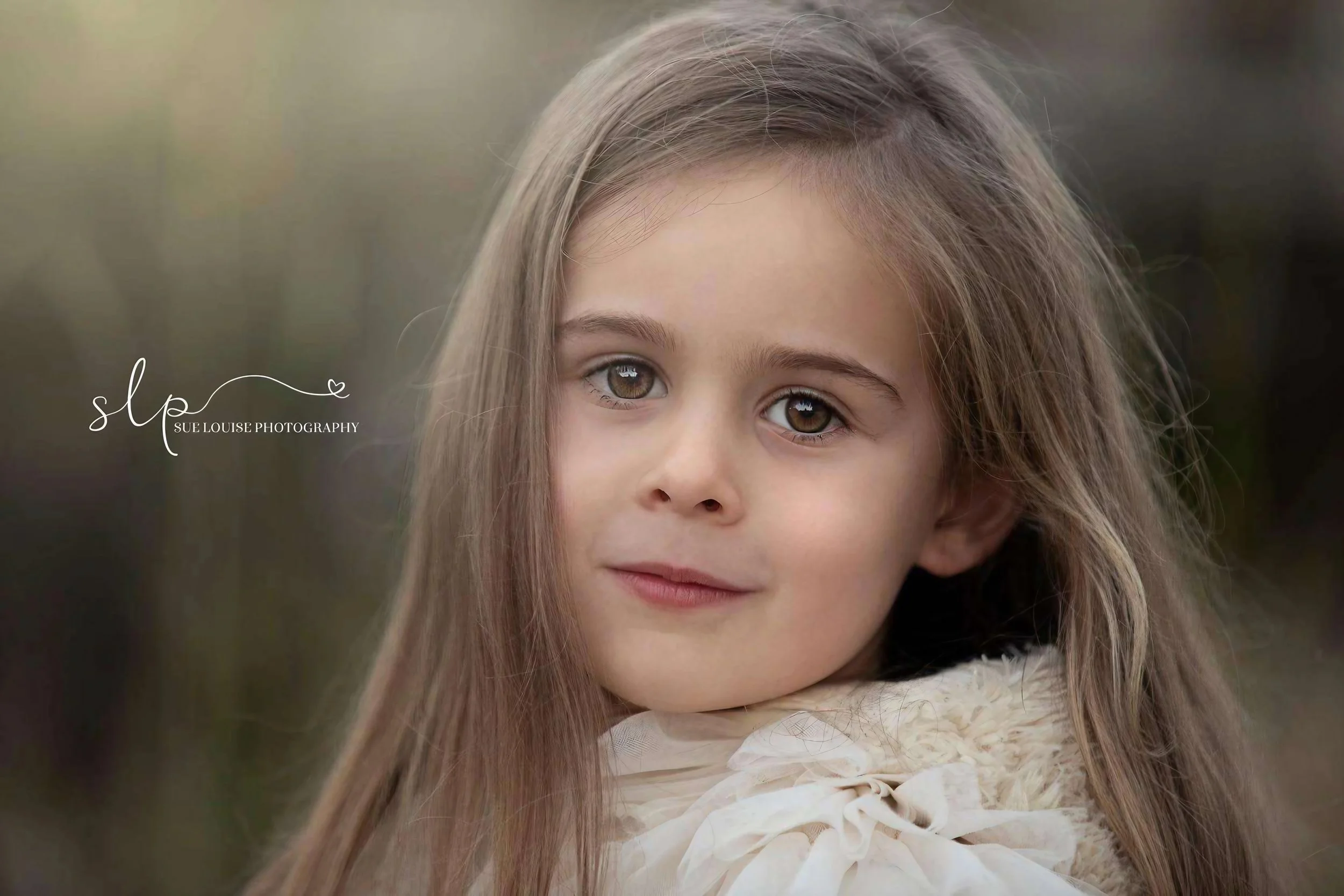 Close-up portrait of a young girl with long brown hair and hazel eyes, wearing a light-colored top with a bow, outdoors with blurred background.