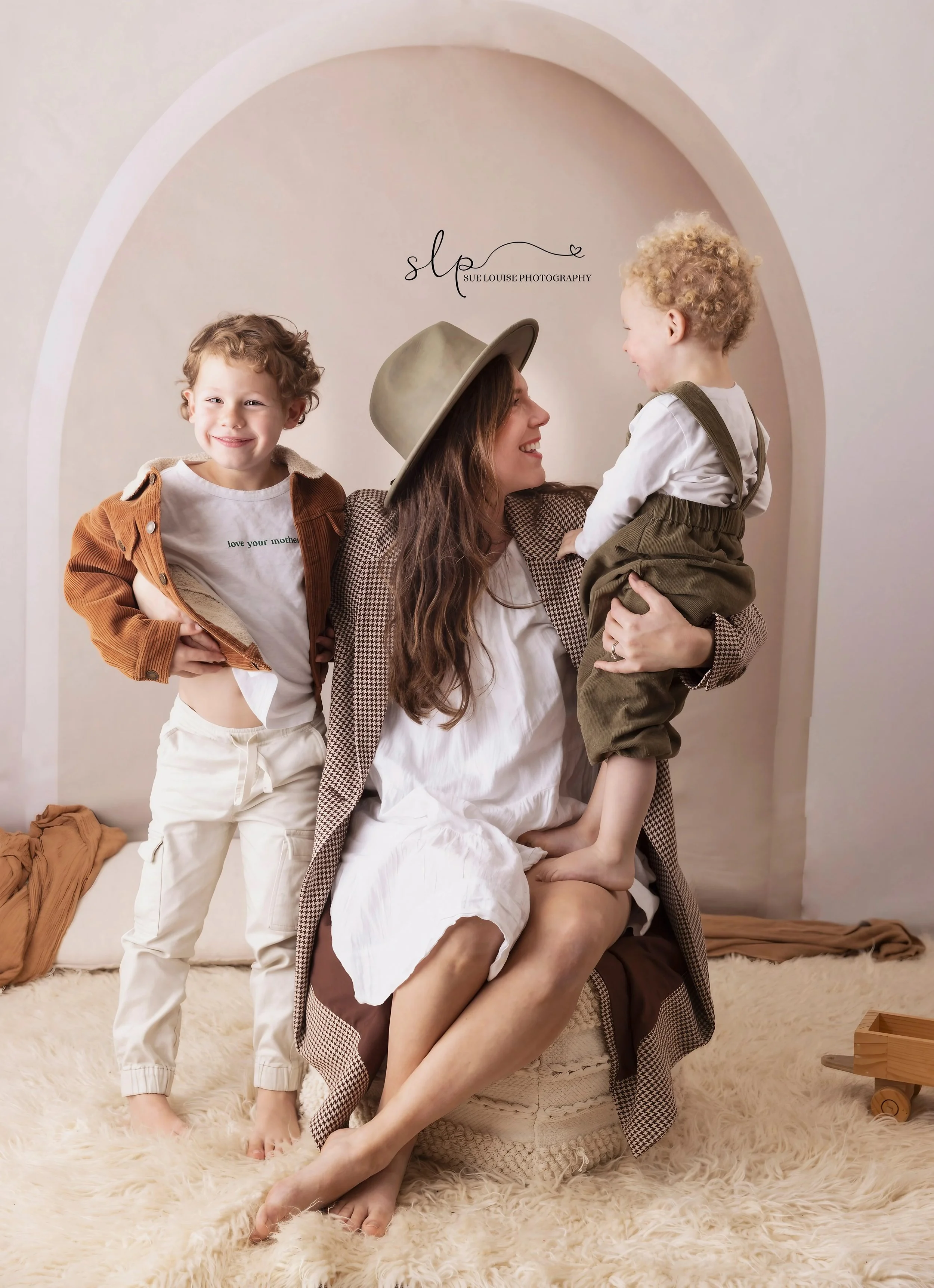 A woman sitting on a cozy, textured pouf in a cream-colored room with two young boys, one standing to her side and one sitting on her lap, all smiling and engaging happily with each other.