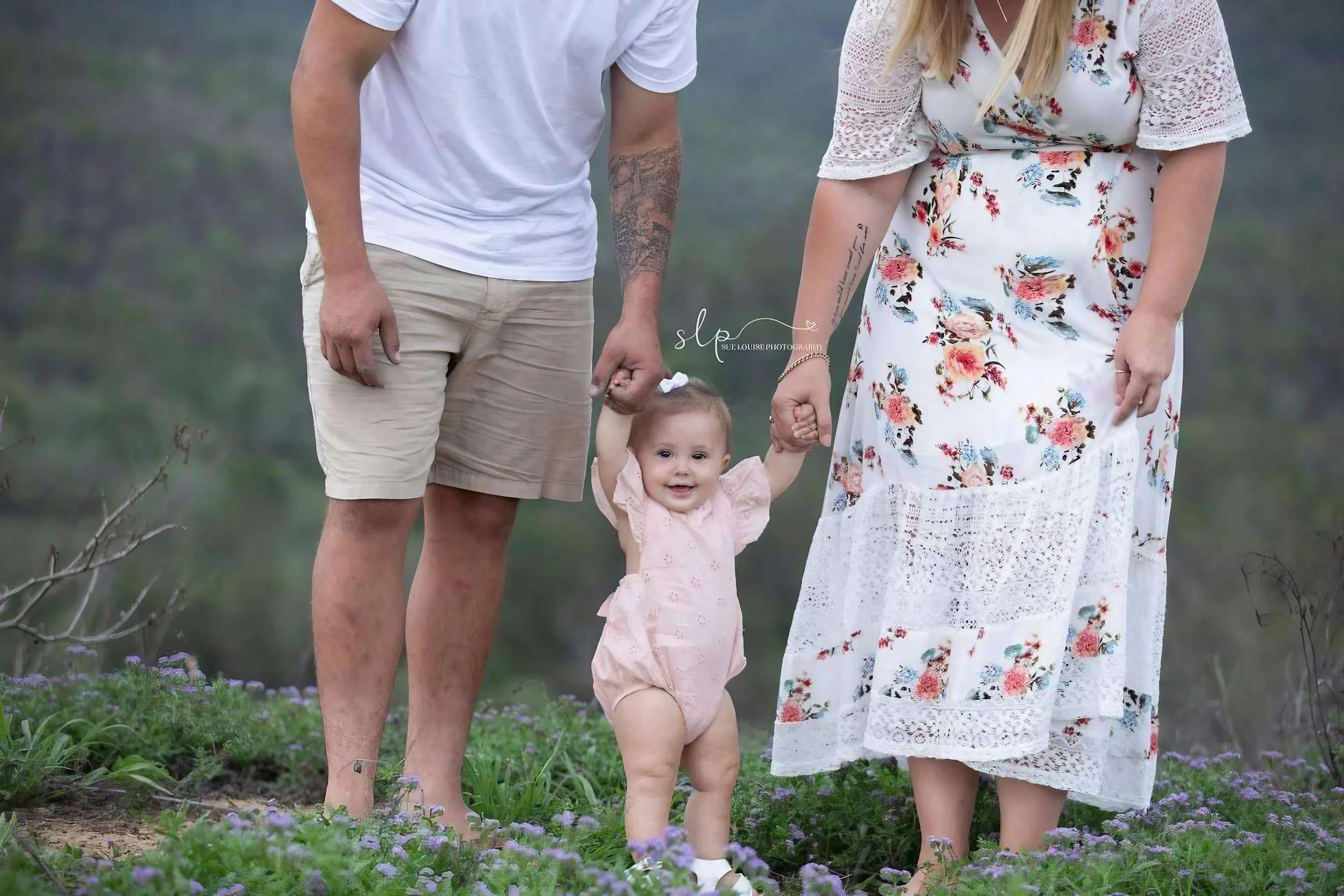 A smiling toddler girl in a pink dress standing between her parents outdoors on purple flowers, holding their hands.