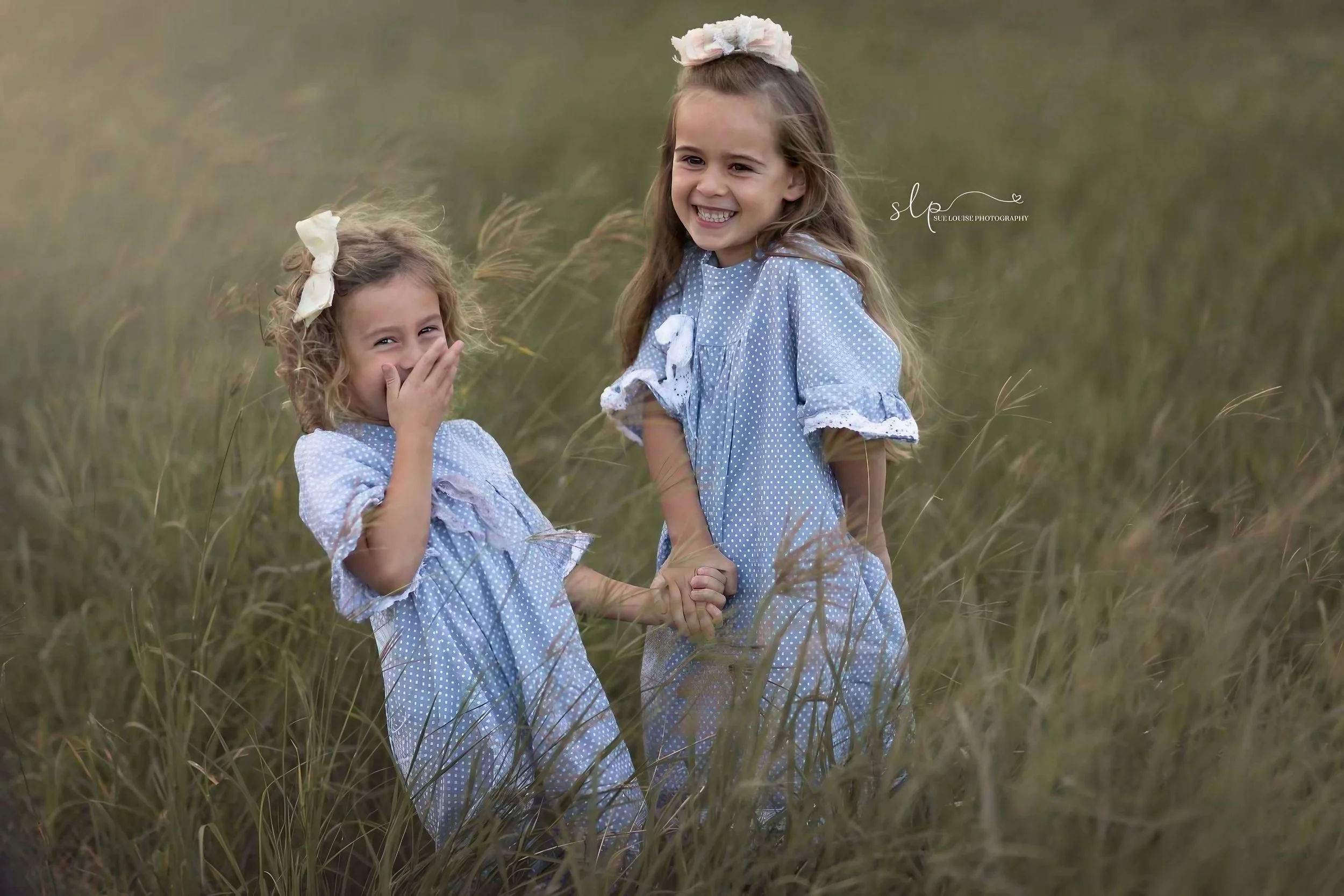 Two young girls in blue polka-dot dresses holding hands and smiling in a grassy field, one covering her mouth in surprise and the other smiling broadly.