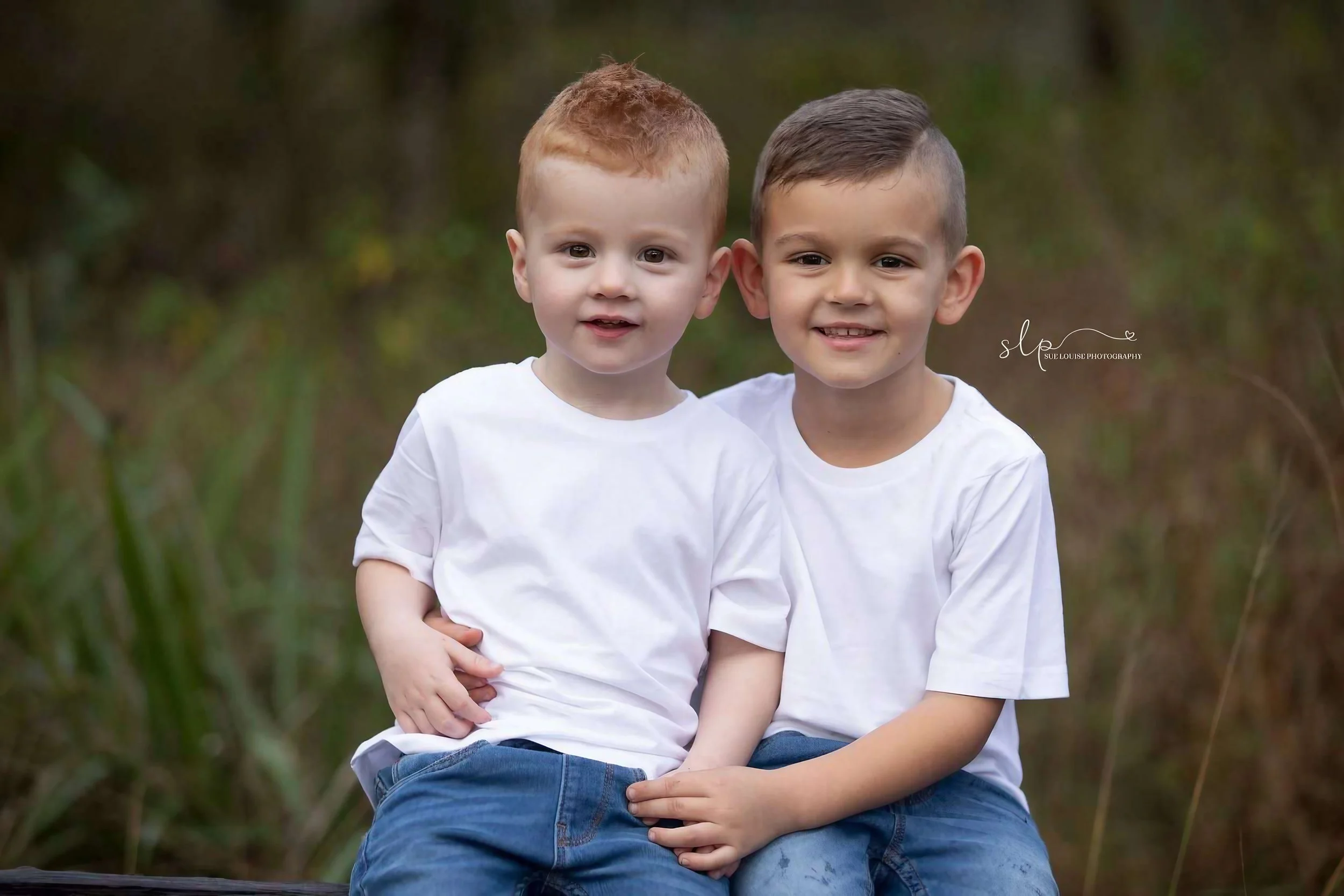 Two young boys sitting outdoors in front of a blurred natural background, wearing matching white t-shirts and blue jeans, smiling at the camera.