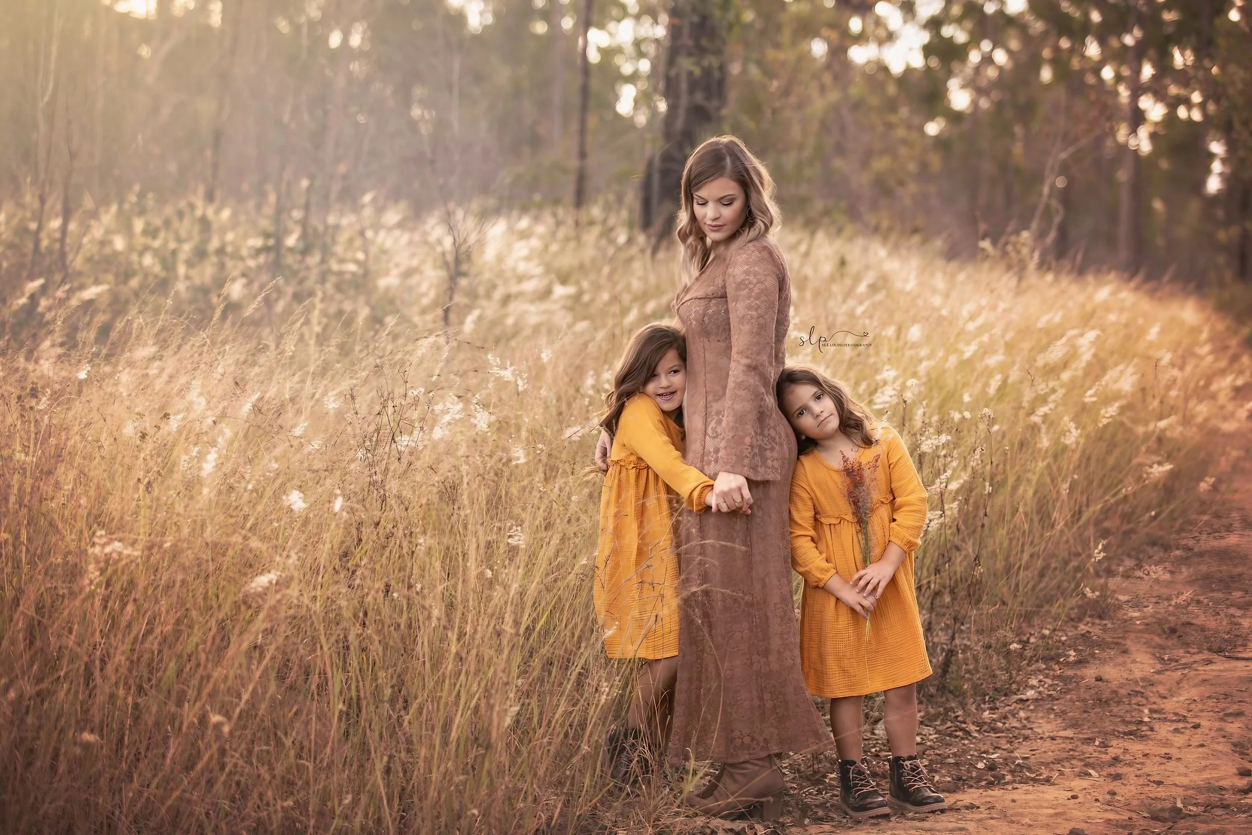 A woman and two young girls standing in a field of tall, golden grass during sunset. The woman, dressed in a brown dress, stands with her eyes closed while hugging one girl who is smiling, and the other girl leans her head on the woman's shoulder, lo