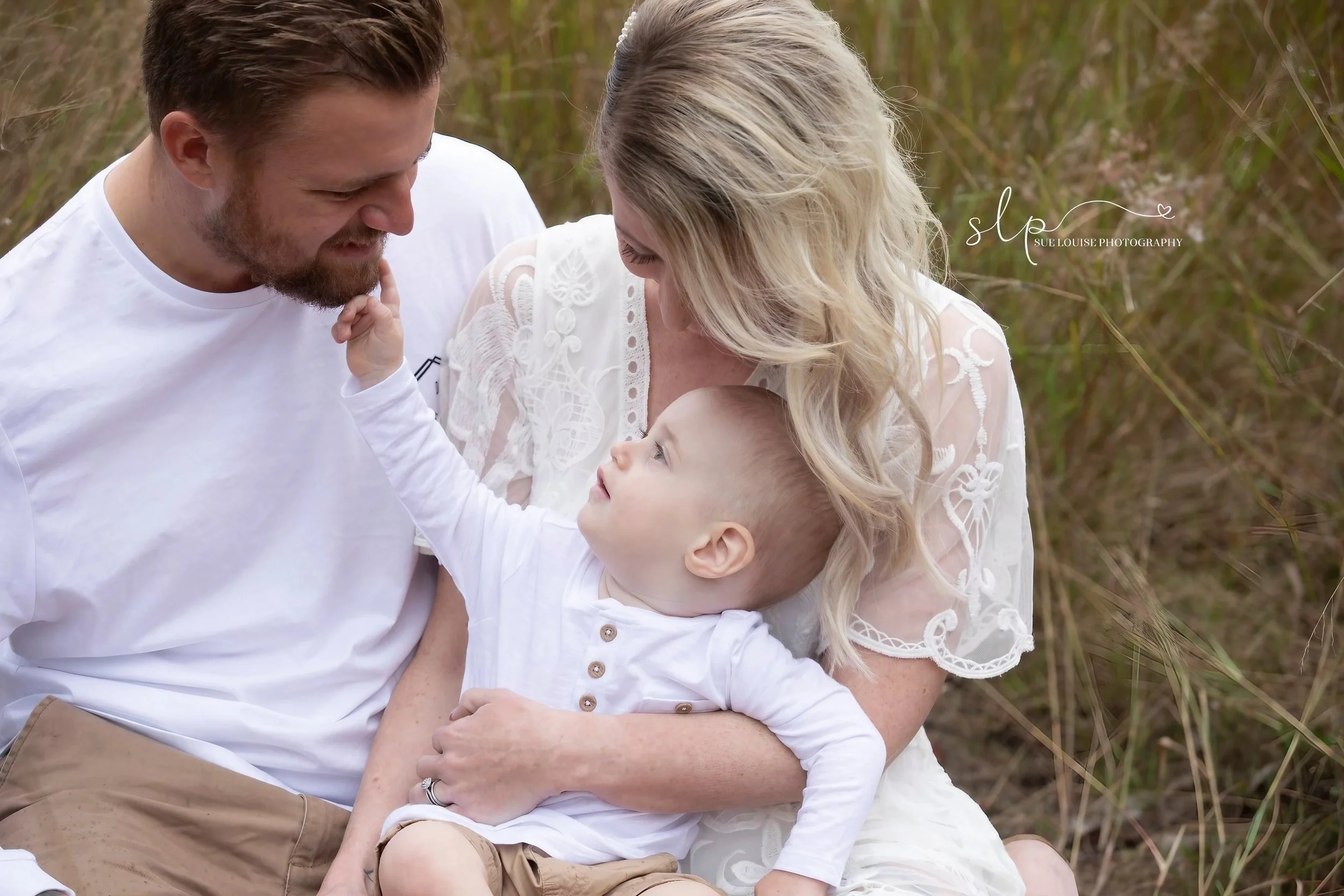 A family of three sitting outdoors in a grassy area, with the father and mother looking at their young son who is sitting between them. The mother is holding the son in her arms, and they are all smiling and enjoying each other's company.
