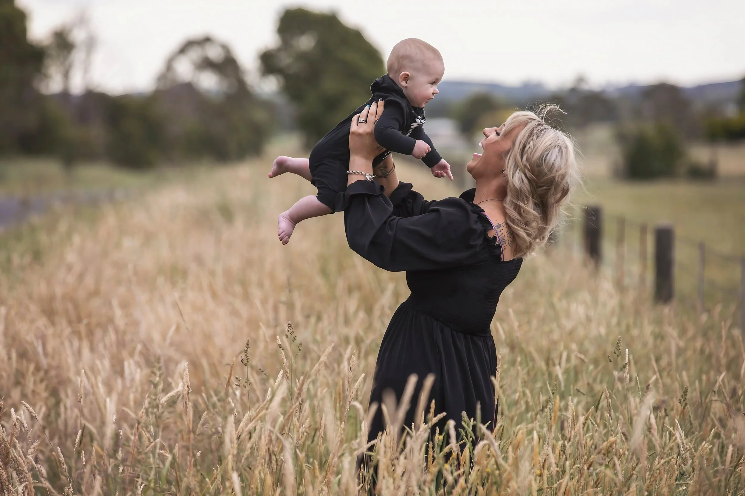 A woman with blonde hair in a black dress holding a smiling baby boy in a field of tall grass, with trees and cloudy sky in the background.