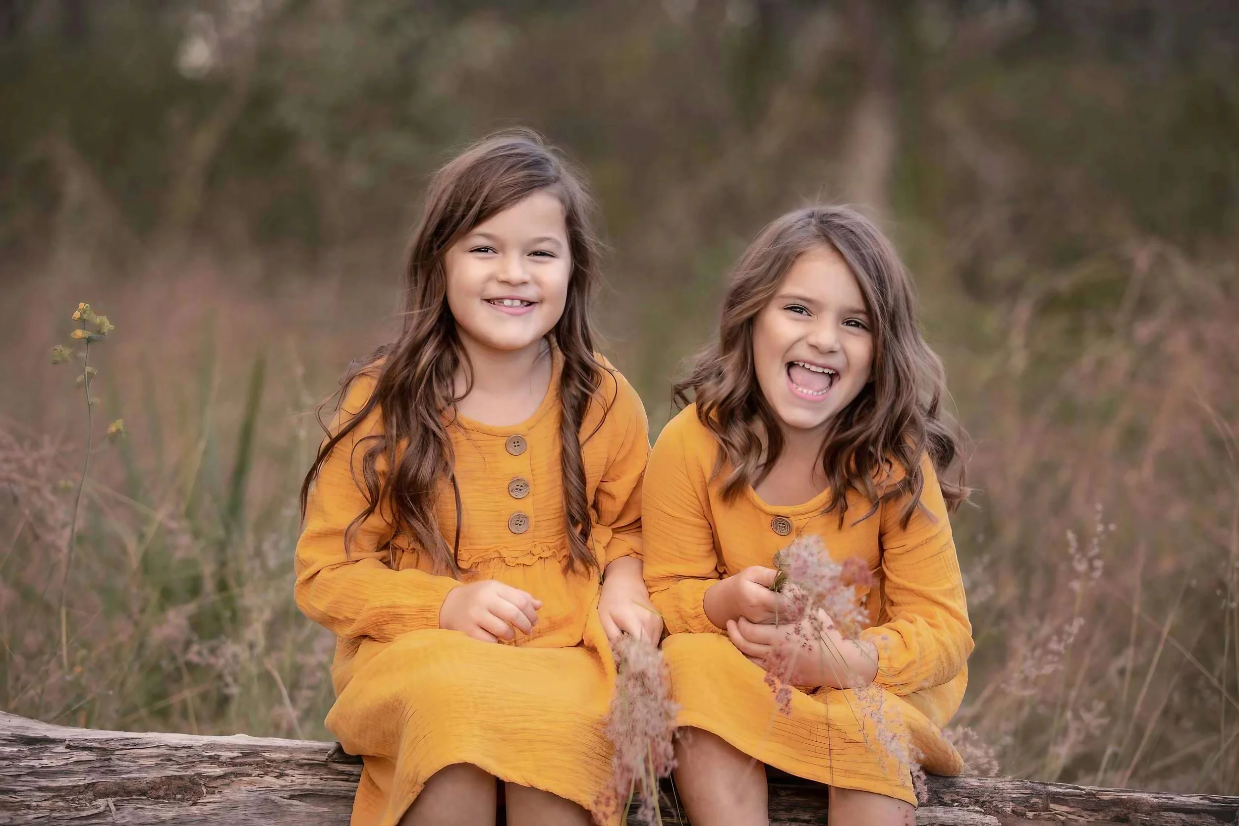 Two young girls with long hair wearing matching yellow dresses, sitting on a log in a natural outdoor setting, smiling and laughing.