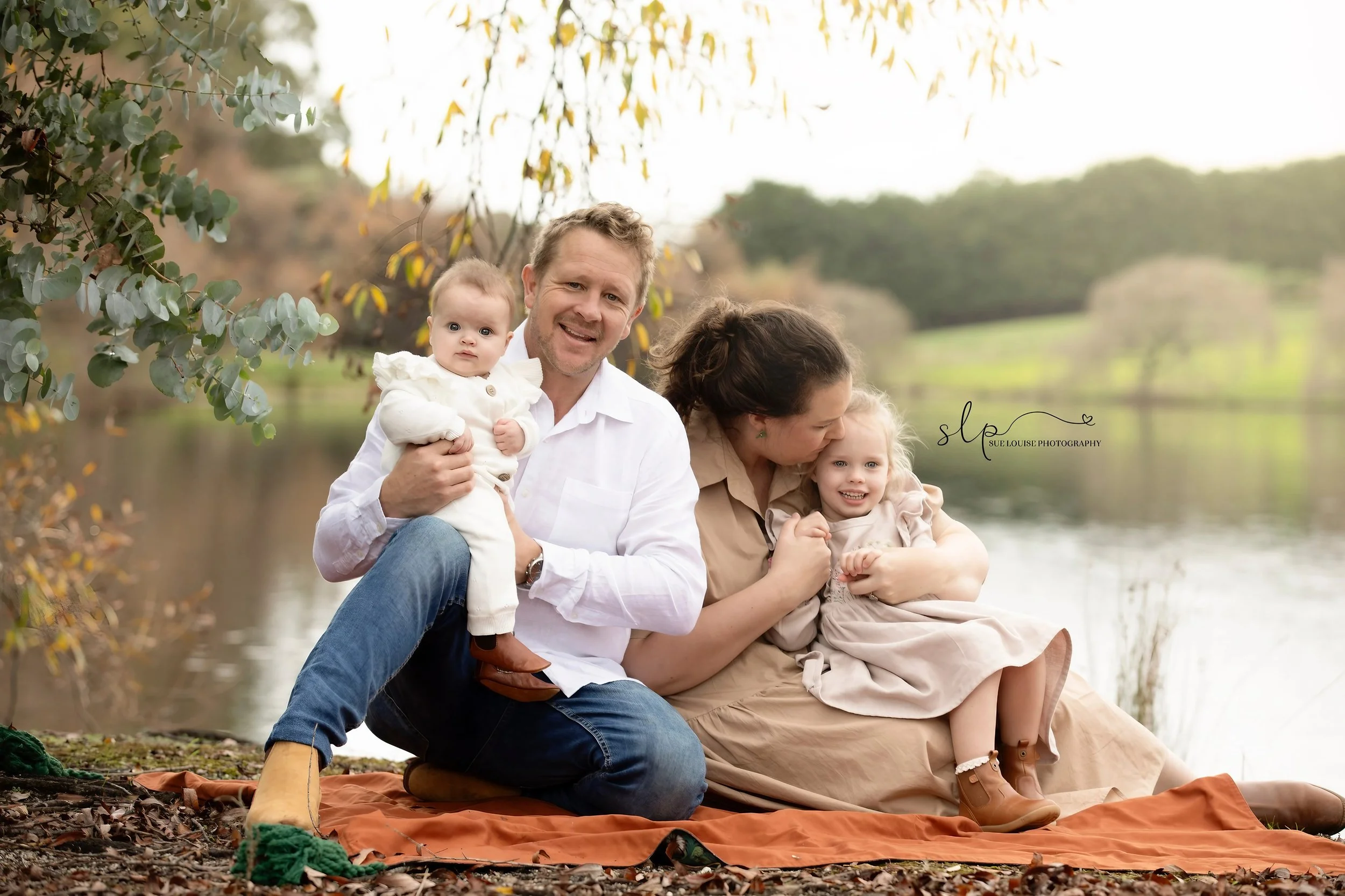 A family of four sitting on a blanket by a lake with trees in the background. The father holds a baby, and the mother is kissing a young girl on the cheek. The family appears happy and enjoying a fall day outdoors.