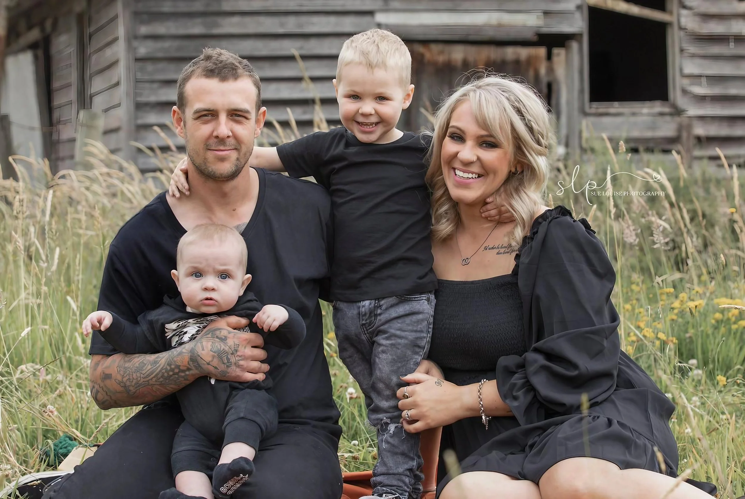 A family of four sitting outdoors in a grassy field with a rustic wooden structure in the background. The father has tattoos and is holding a baby, the mother is smiling and has blonde hair, and a young boy is sitting on the father's shoulder with hi