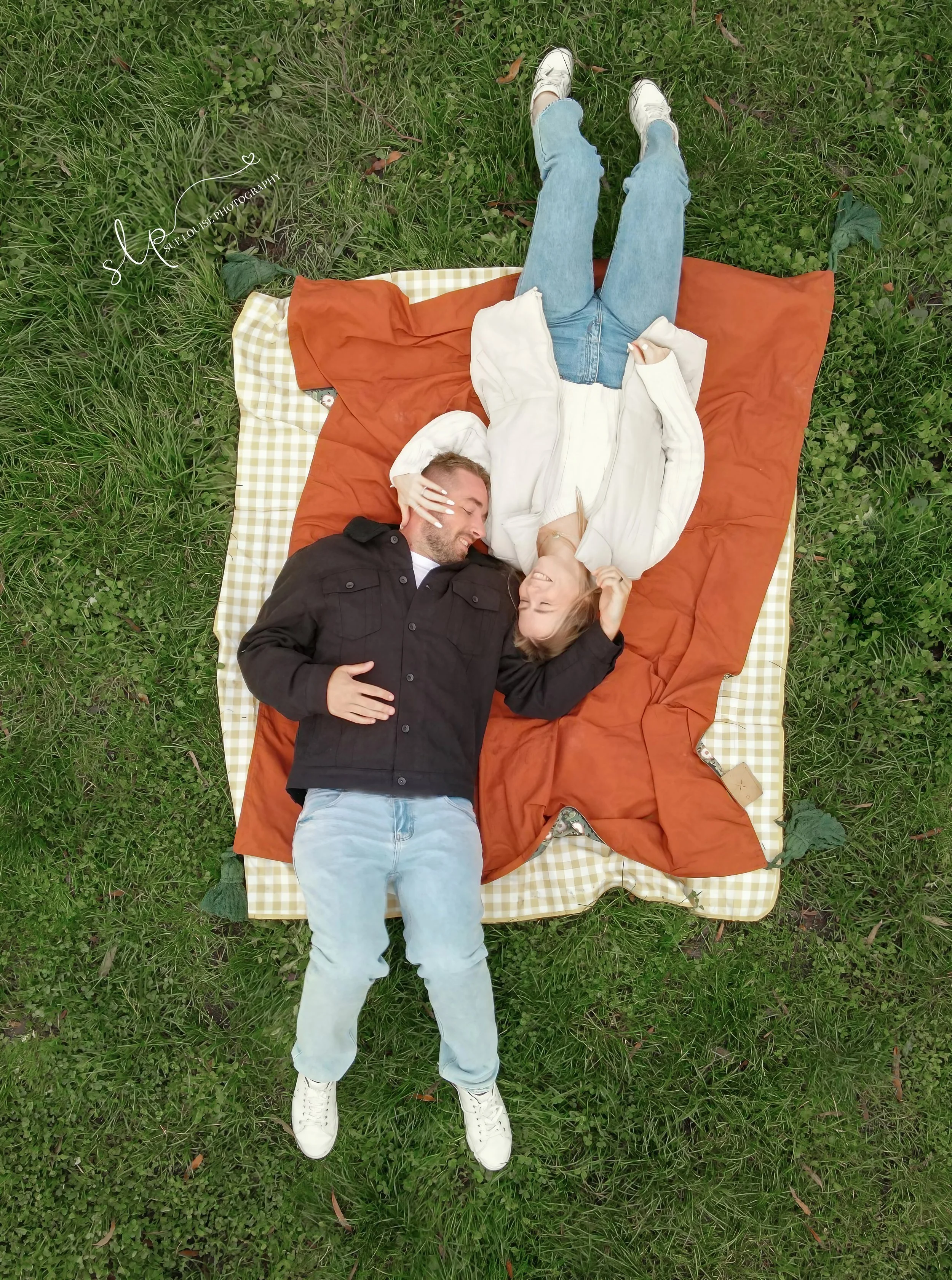 A man and woman lying on a blanket outdoors on the grass, facing each other and smiling. The woman is wearing a white jacket and jeans, and the man is wearing a black jacket and light jeans.