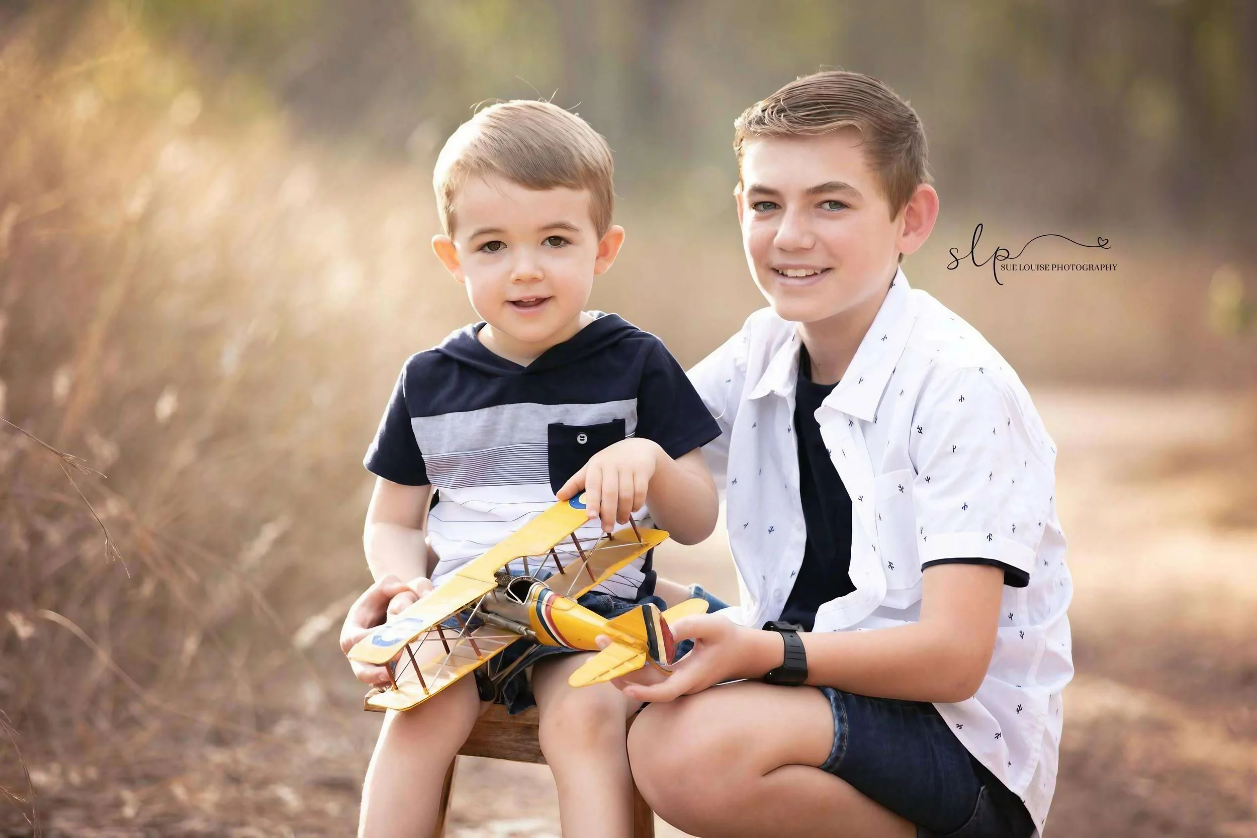 A young boy and a slightly older boy sitting outdoors holding a yellow and black toy airplane, smiling at the camera, with a natural background of blurred trees and foliage.