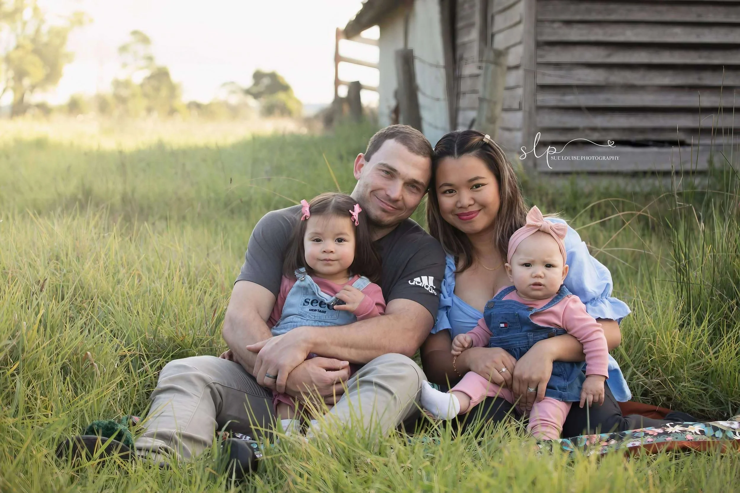 Young family of four sitting on grass near a barn, smiling at the camera, with the father and mother holding their two daughters.