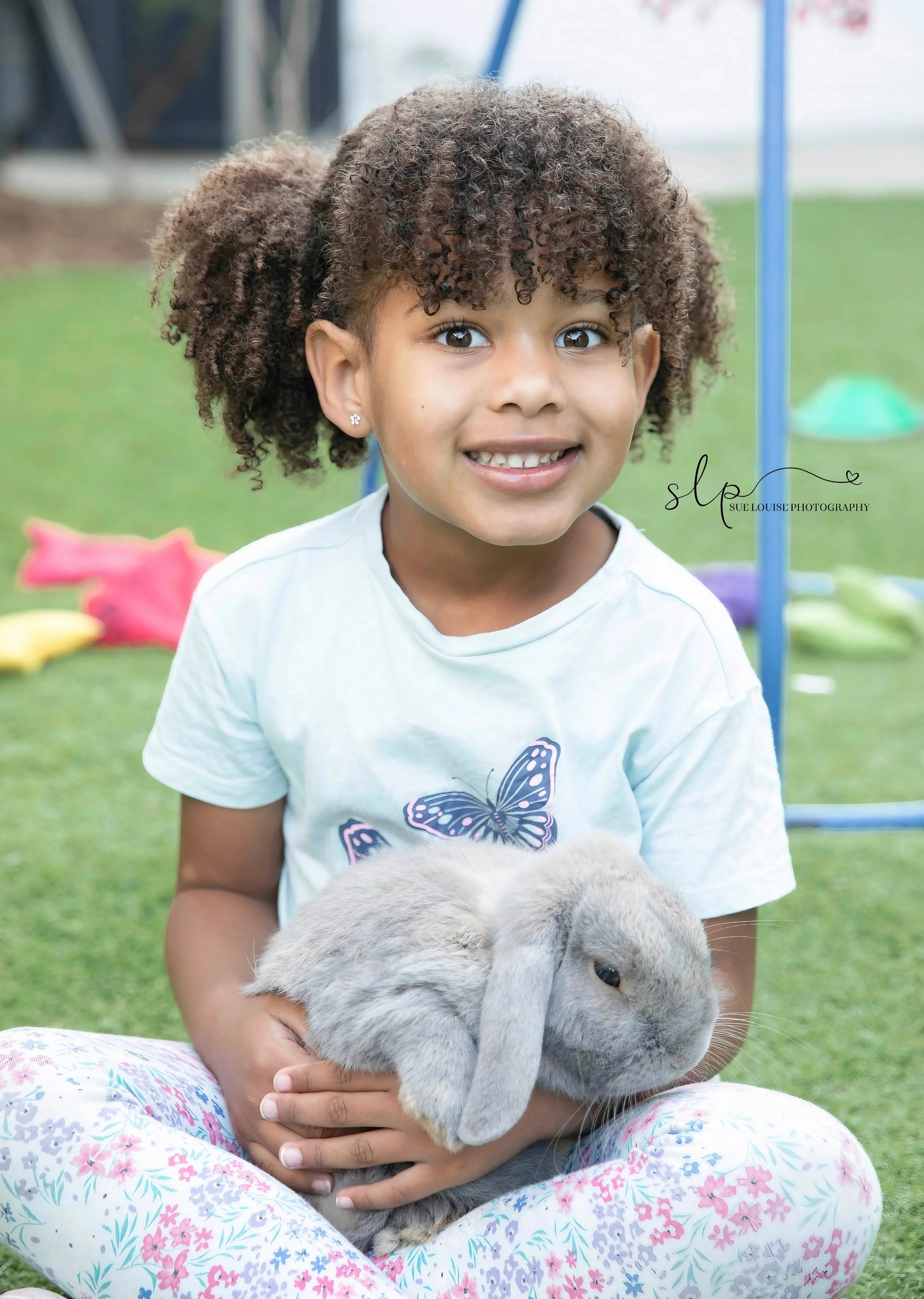 A young girl with curly brown hair wearing a white t-shirt with a butterfly design and floral pants, sitting on grass and holding a gray rabbit. She is smiling at the camera. Background includes playground equipment and colorful toys.