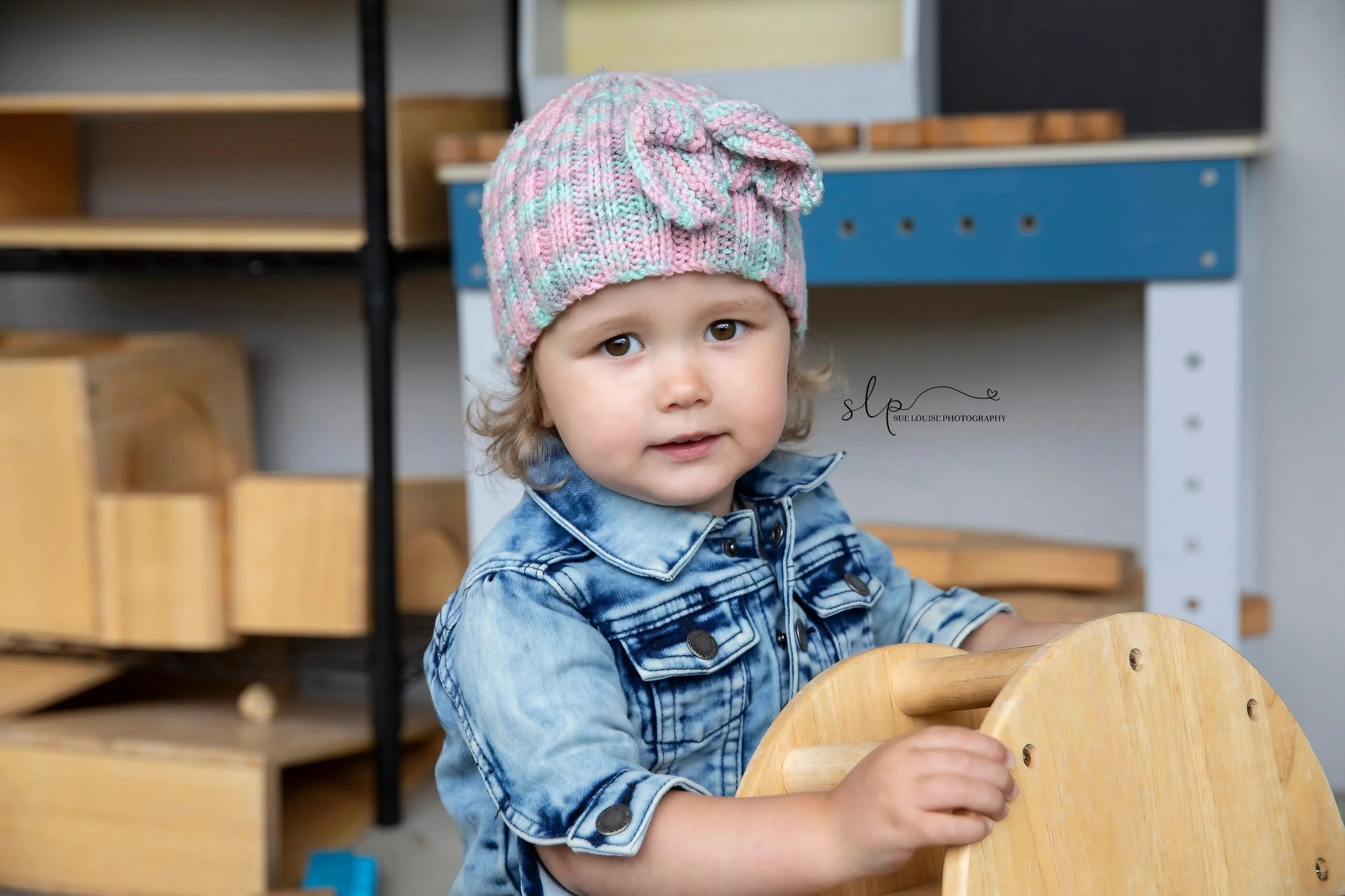 A young girl with curly hair wearing a colorful knit hat with a bow and a denim shirt, sitting at a wooden chair in a playroom or classroom setting.