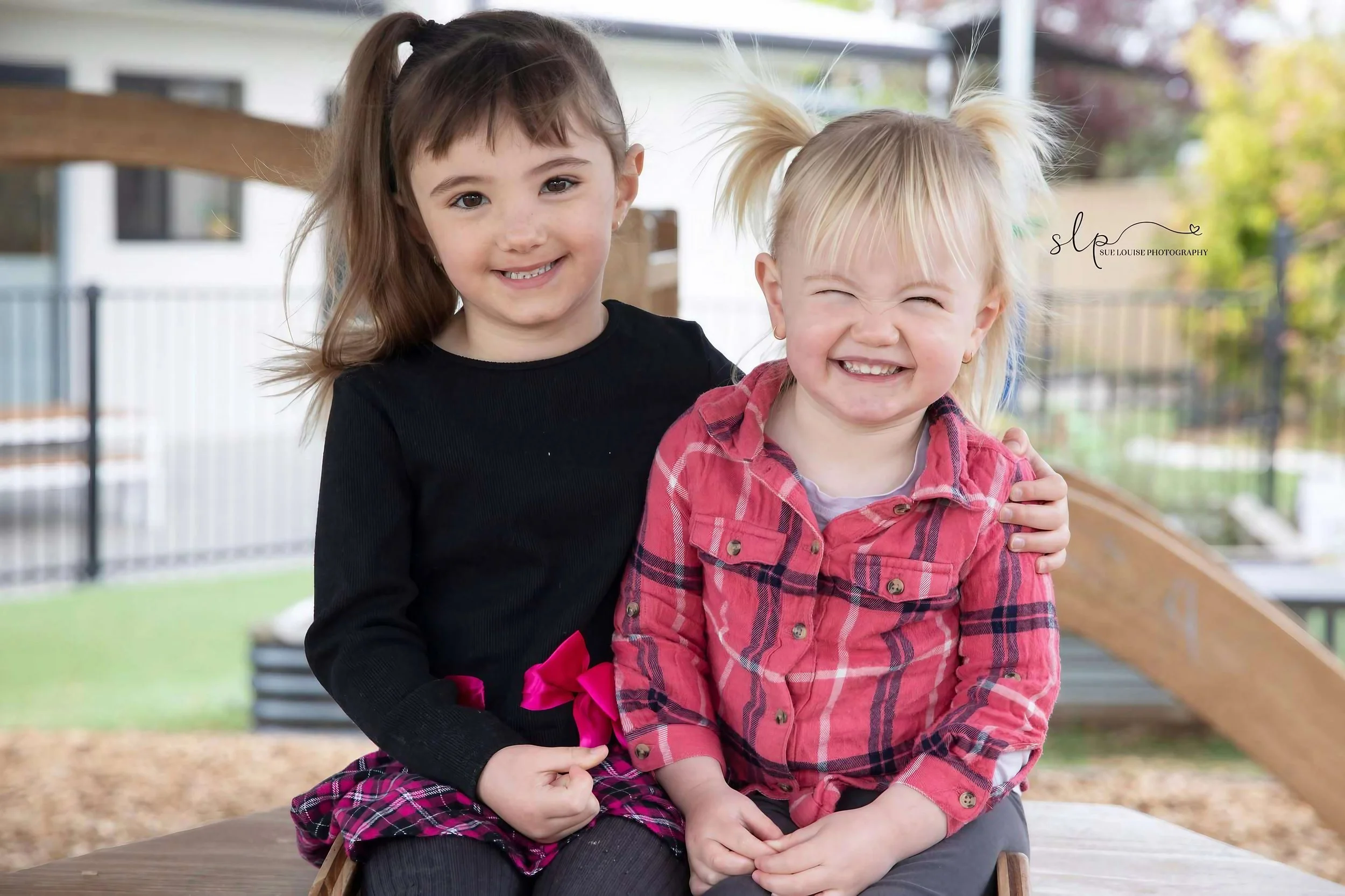 Two young girls sitting outdoors, smiling and happy. One girl has brown hair in a ponytail, wearing a black shirt and plaid skirt with a pink bow. The other girl has blonde hair in pigtails, wearing a red plaid shirt and gray pants. They are sitting