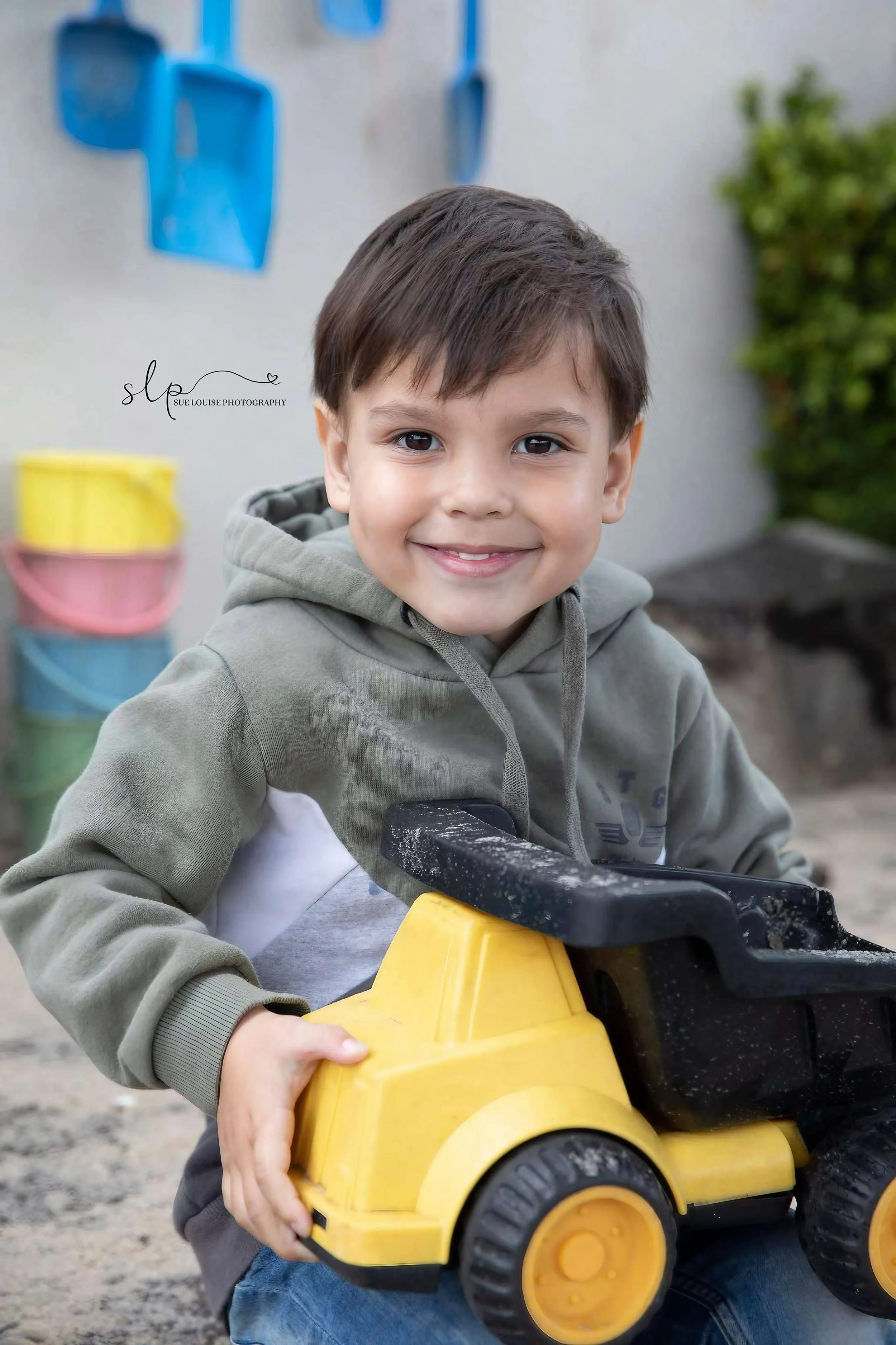 A young boy smiling and holding a yellow plastic toy bulldozer at a playground with colorful buckets in the background.