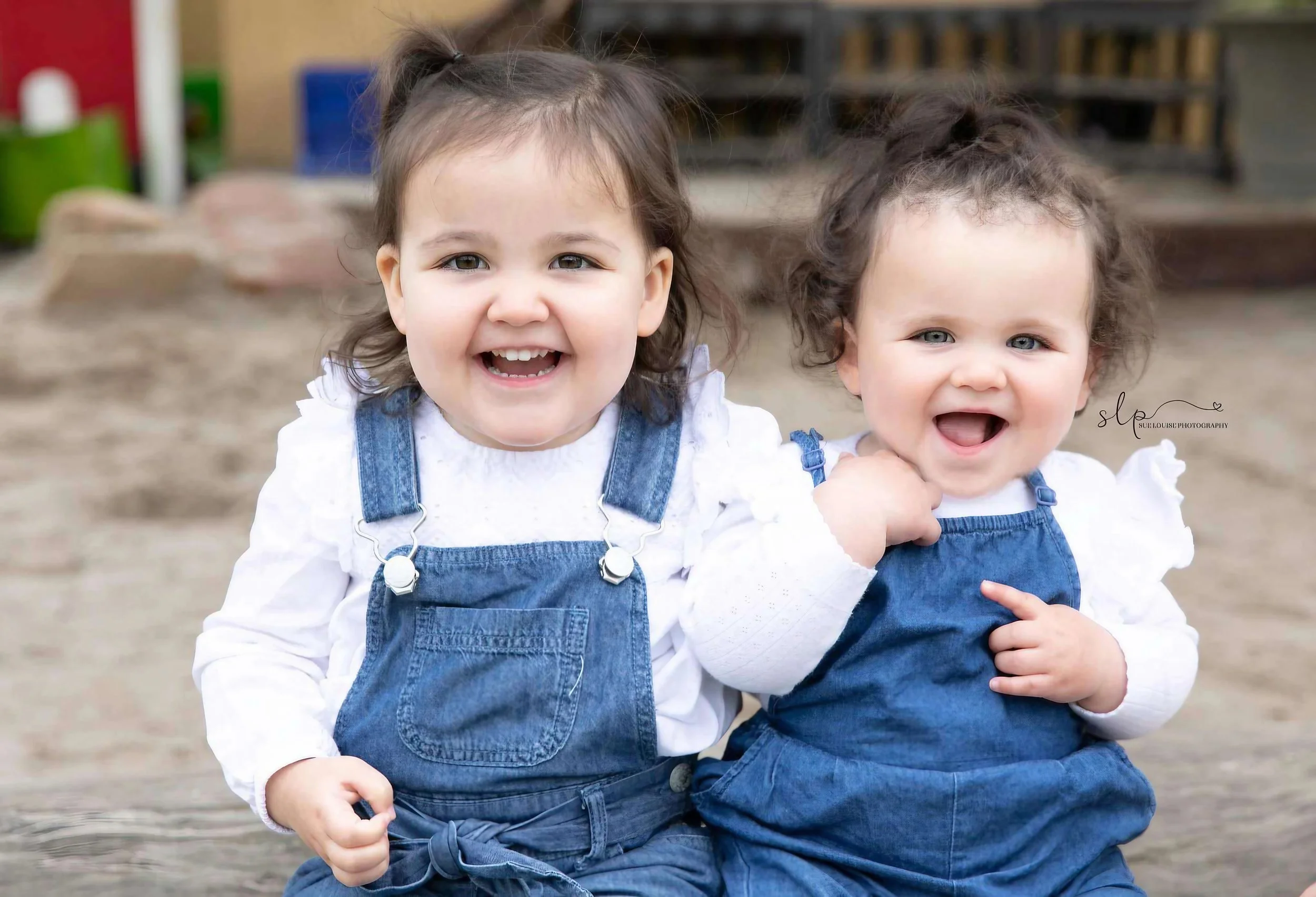 Two young girls with curly hair smiling and sitting on a wooden surface outdoors, wearing blue denim overalls and white shirts.