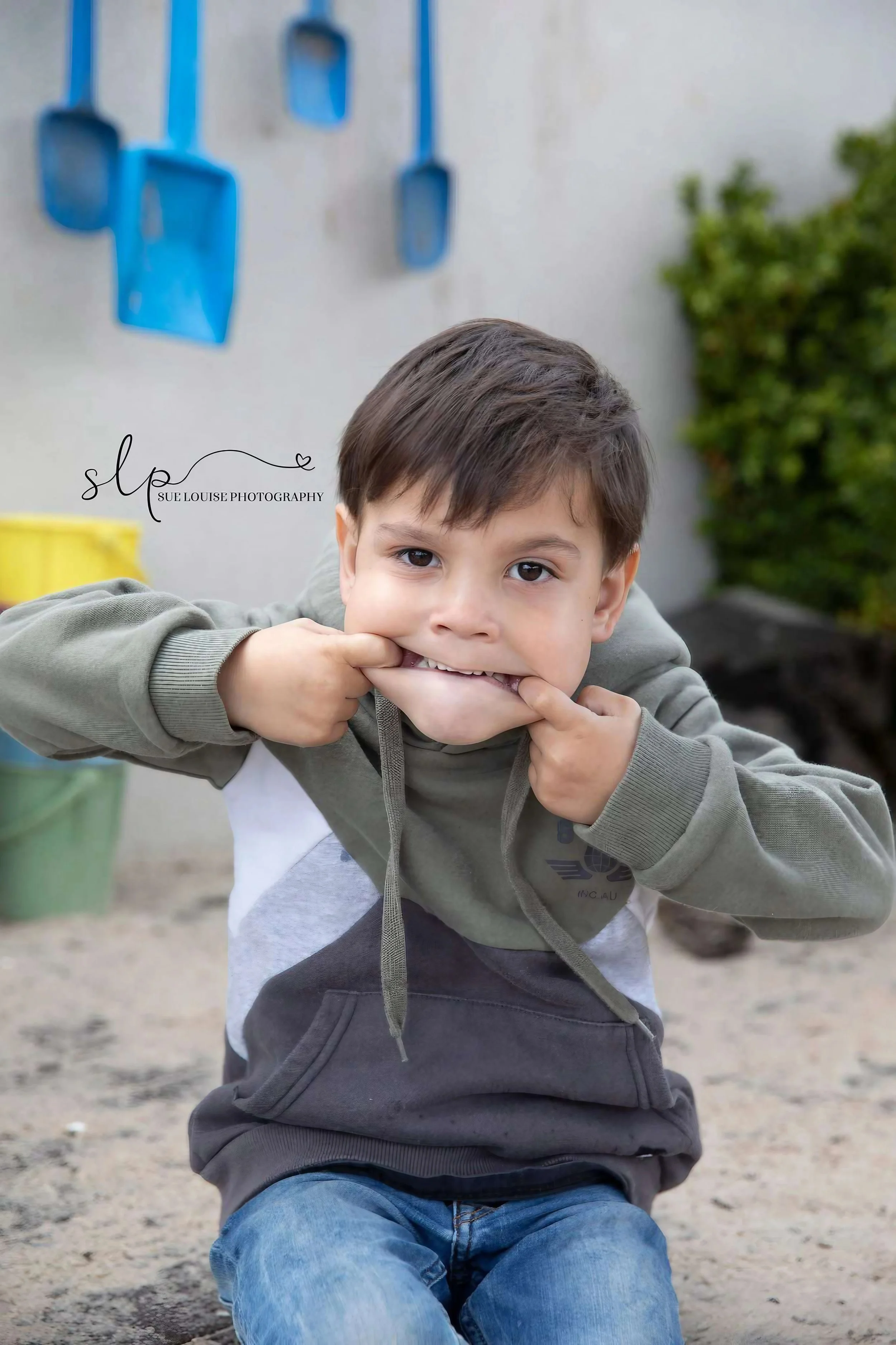 Young boy sitting outdoors, pulling his bottom lip with his fingers, making a playful face. He has brown hair and is wearing a gray and white hoodie and blue jeans. In the background, there are colorful planters attached to a white wall and some gree