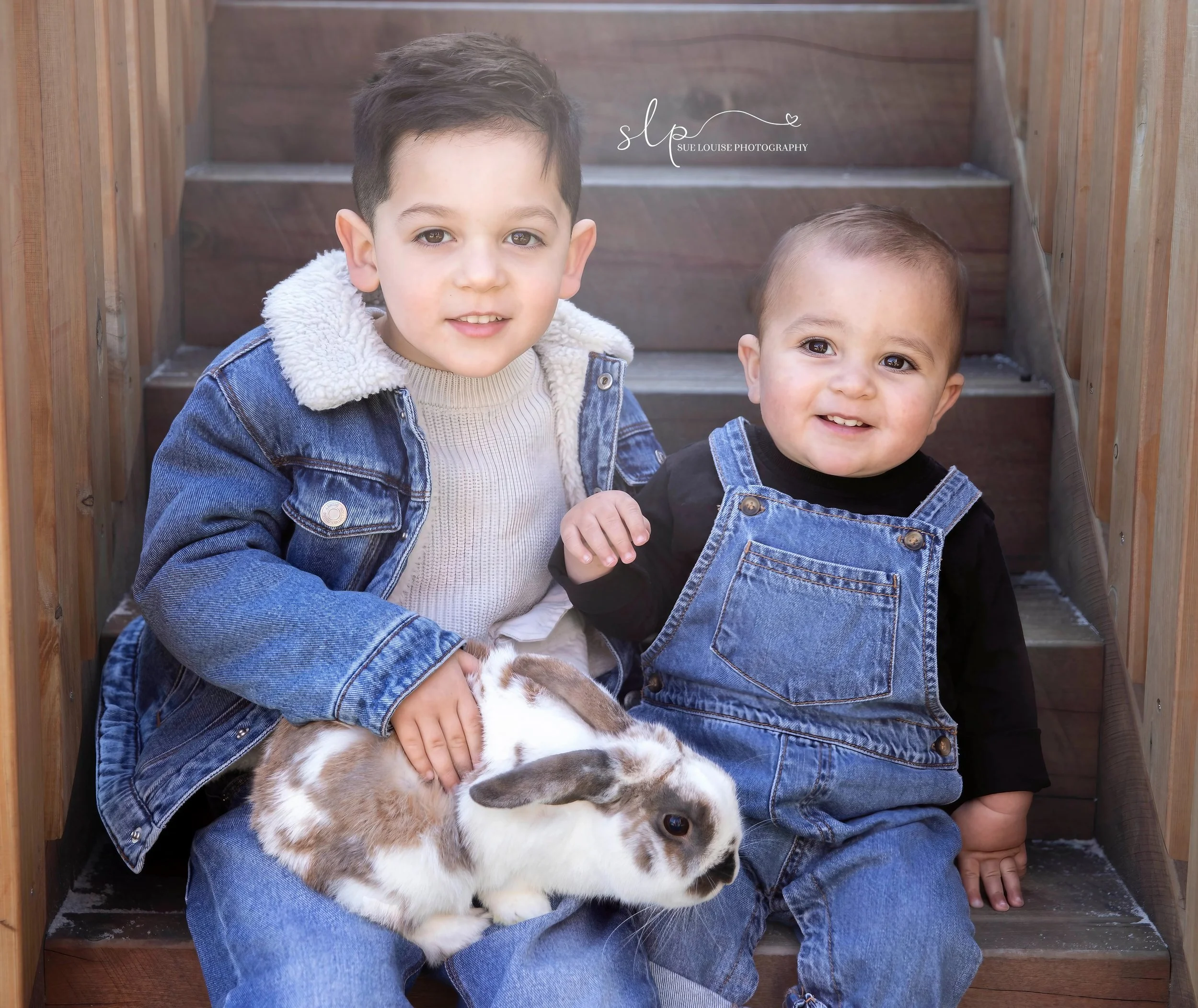 Two young boys sitting on outdoor wooden steps, holding a rabbit. Both boys are wearing denim outfits and are smiling at the camera.