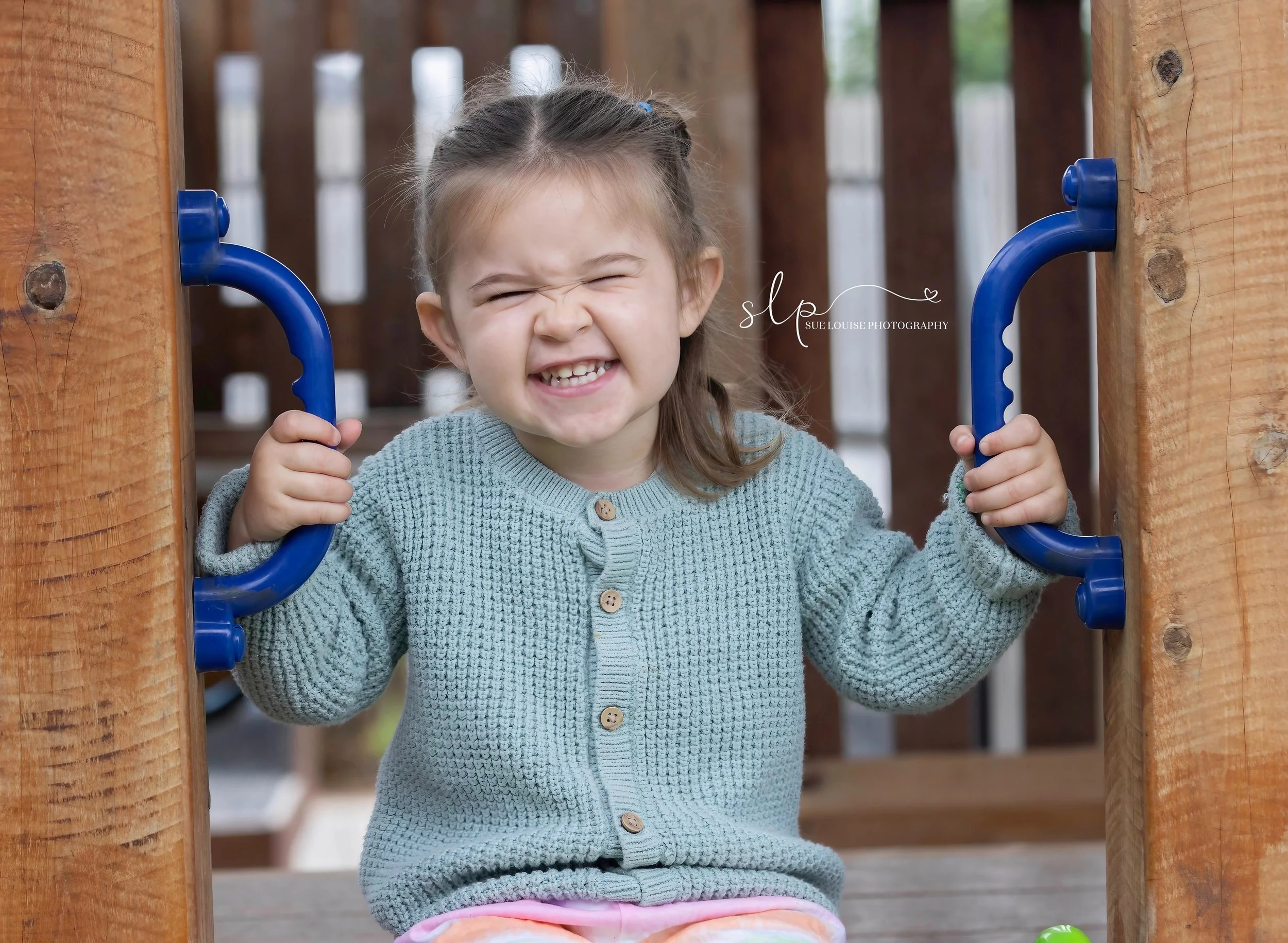A young girl with a gray sweater smiling and squinting her eyes while holding onto a blue play structure on a wooden playground.