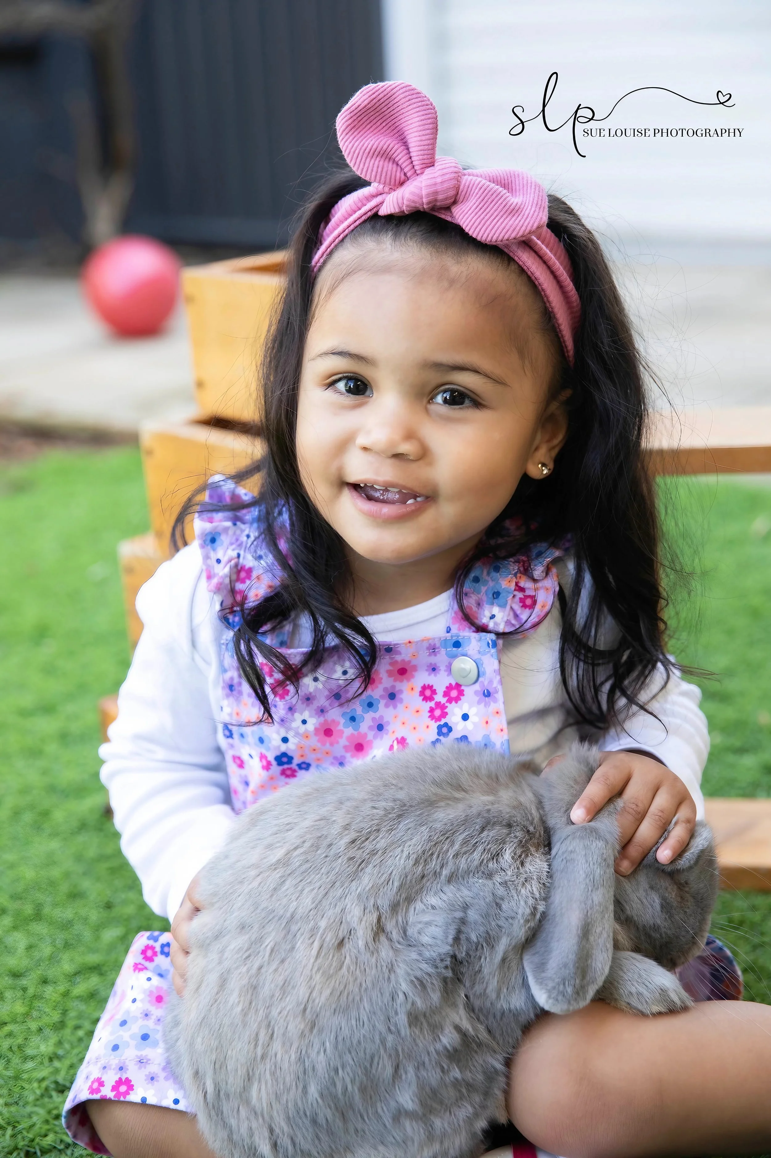 A young girl with long dark hair, wearing a pink headband and a colorful floral dress, sitting outdoors on grass and holding a gray rabbit.