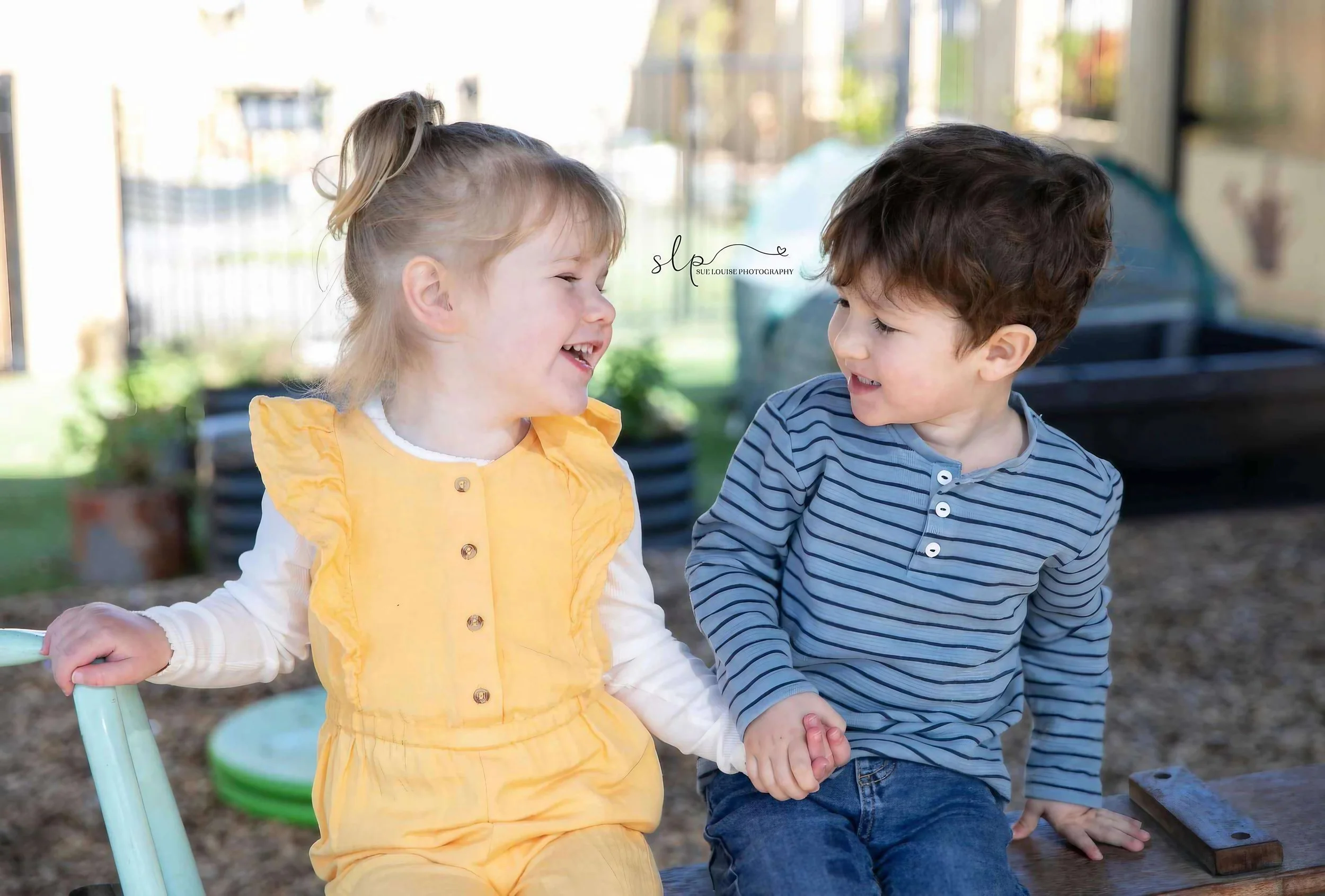 A young girl with blonde hair in a ponytail and wearing a yellow dress, and a young boy with dark hair wearing a blue striped shirt, are sitting outdoors on a bench, holding hands, and smiling at each other.
