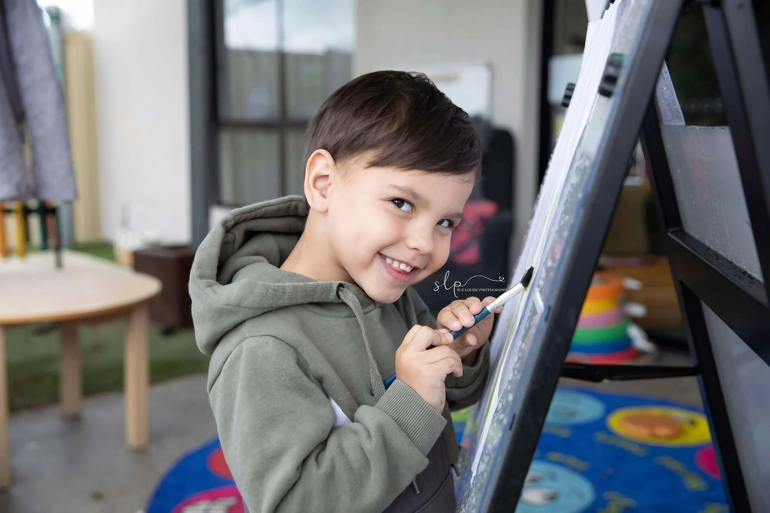 Smiling young boy with short brown hair, wearing a gray hoodie, painting on an easel outdoors.