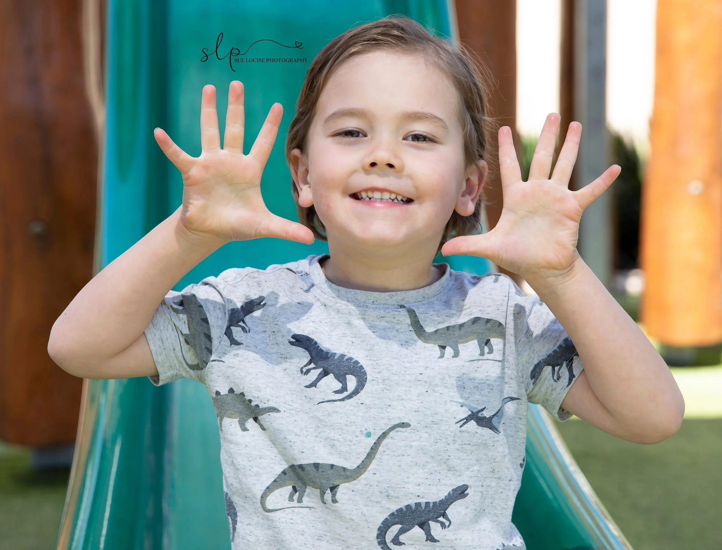 A young boy smiling and holding his hands up near his face, standing outdoors near a slide at a playground.