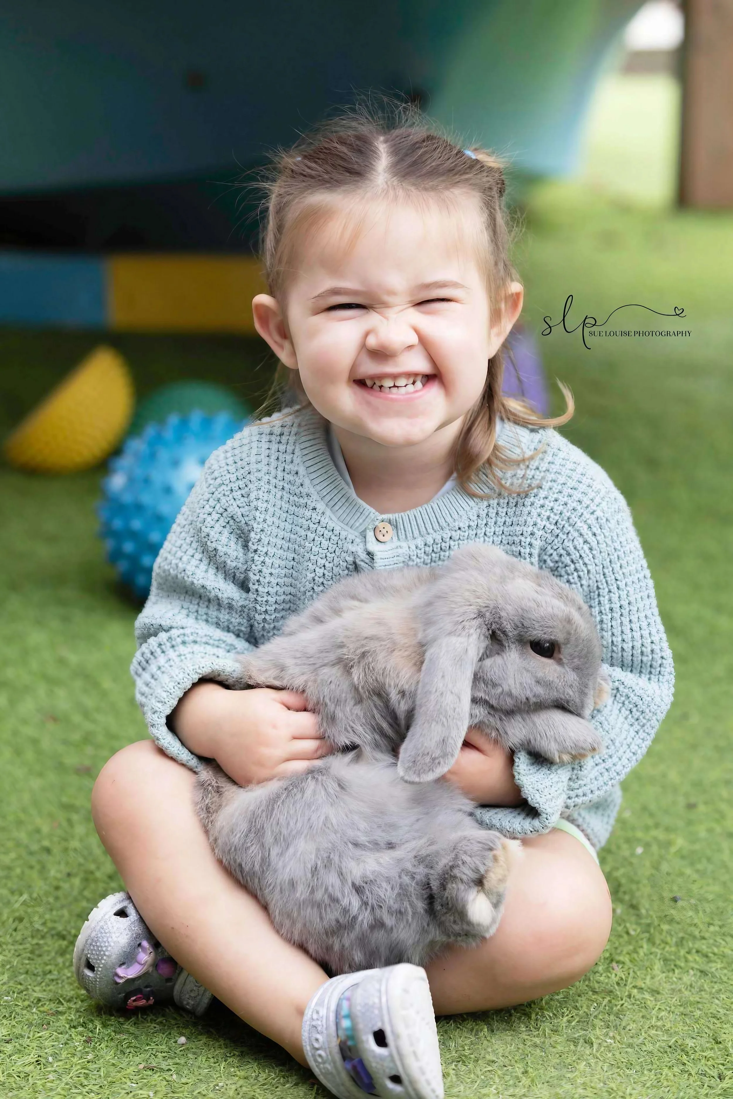 A young girl with a joyful, mischievous smile holding a gray lop-eared rabbit, sitting on green grass with colorful toys in the background.