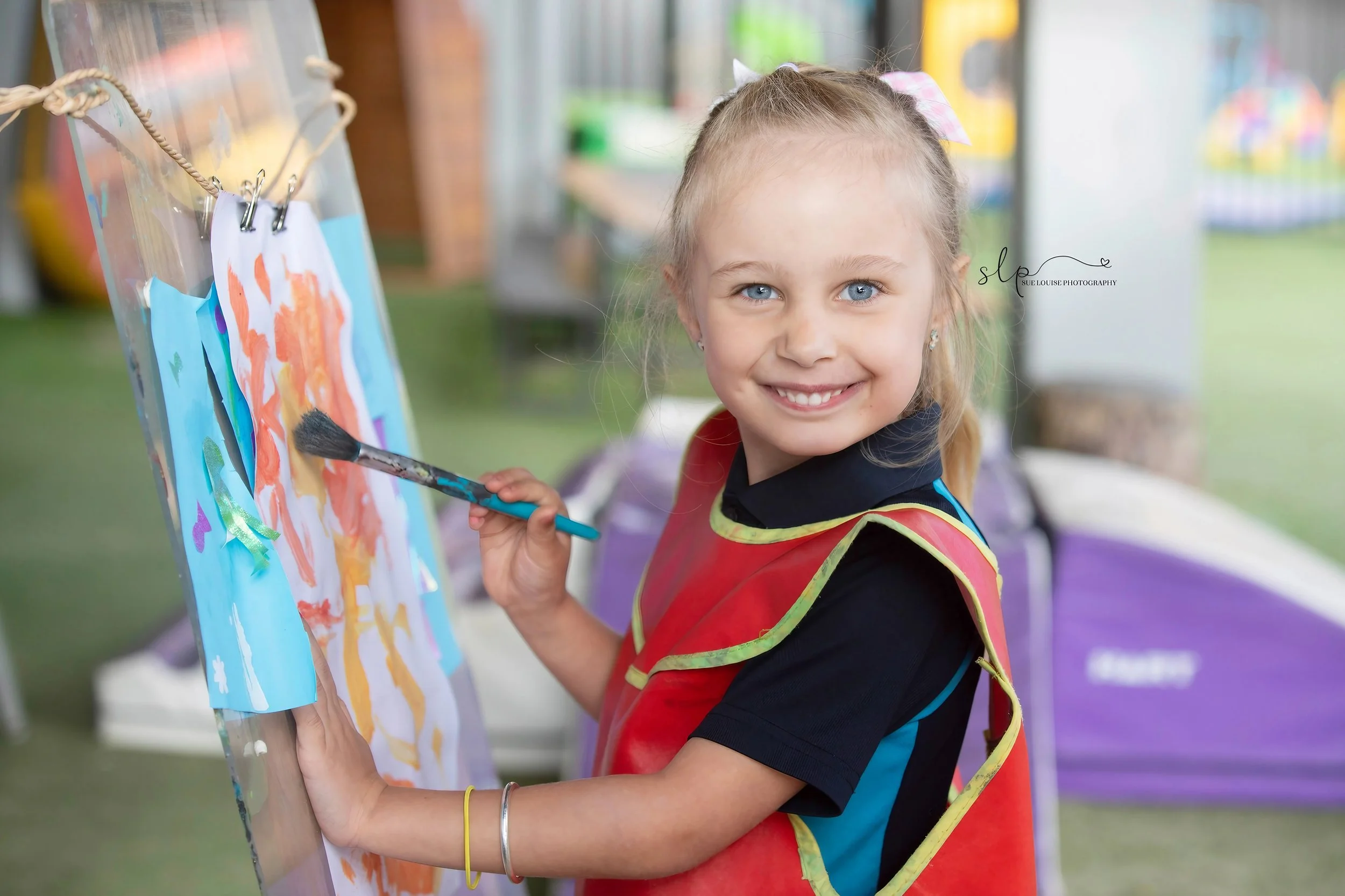 A young girl with blonde hair and blue eyes smiling while painting on an art easel outdoors, wearing a red apron over a black shirt with a pink bow in her hair.