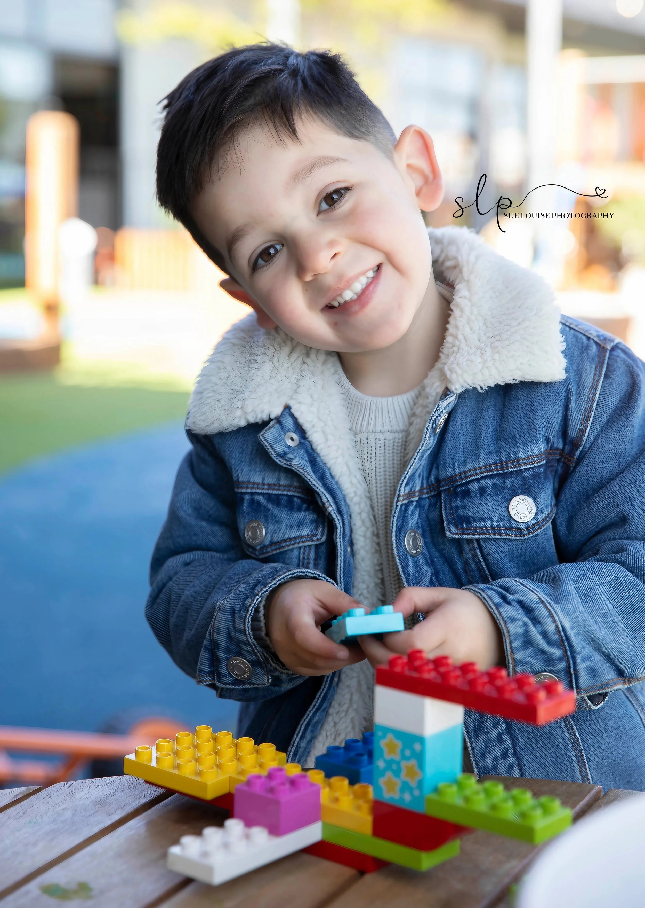 A young boy wearing a denim jacket with a white fleece collar, smiling and playing with colorful building blocks outdoors on a wooden table.