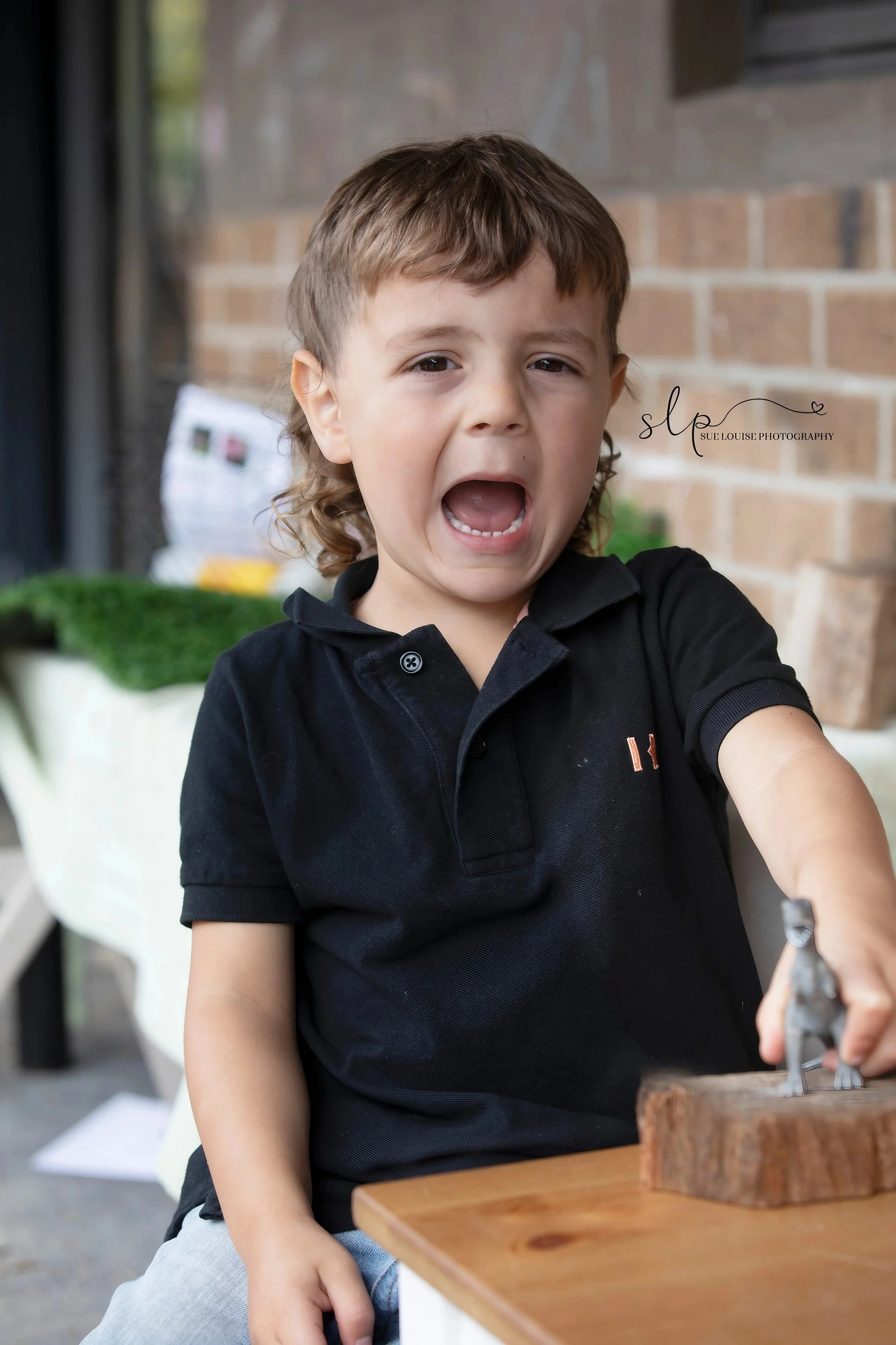 A young boy with brown hair and wearing a black polo shirt is crying and appears upset, reaching out with his right hand towards a small animal figurine on a wooden surface.