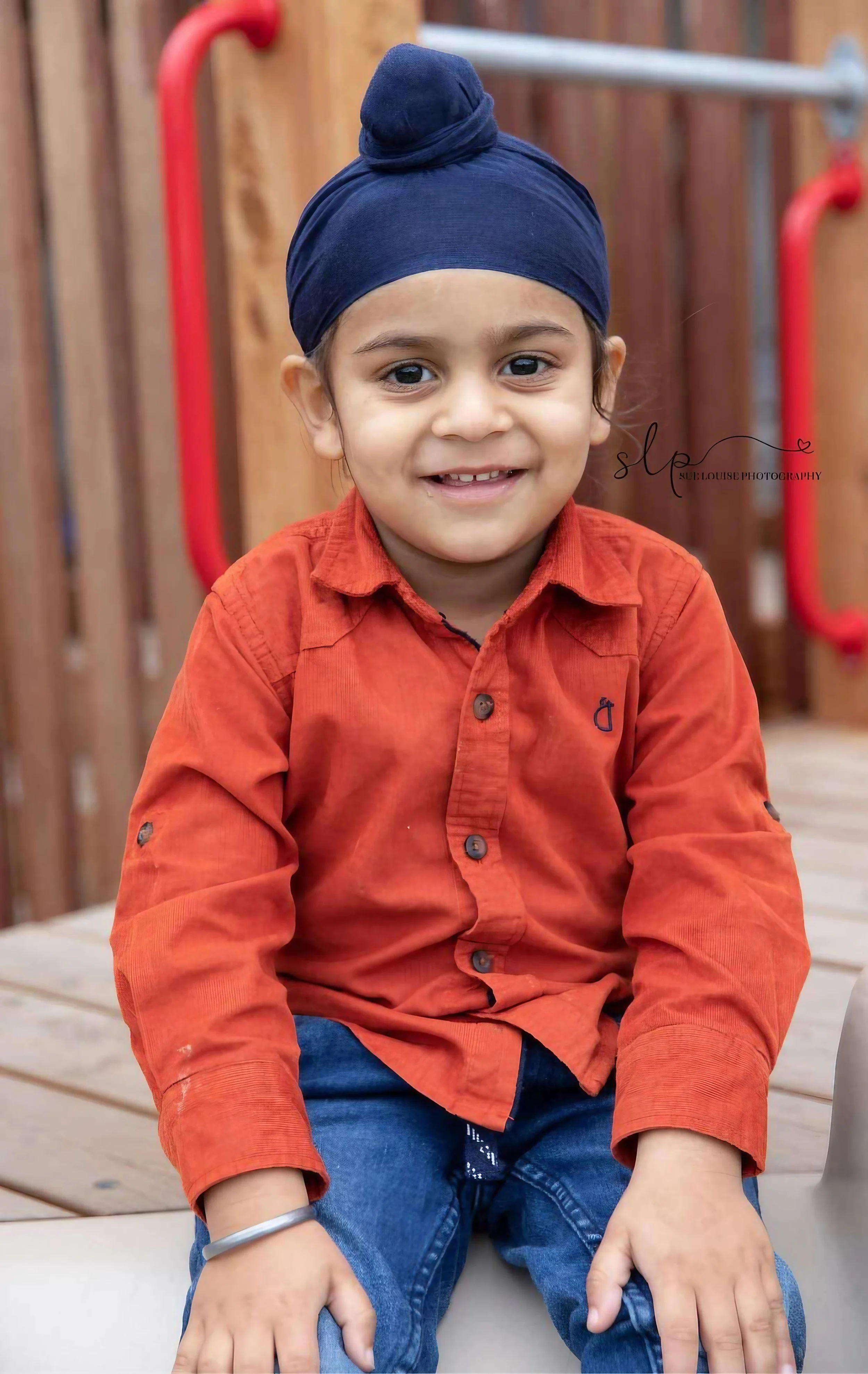 A young boy with dark hair wearing a blue turban, an orange button-up shirt, and blue jeans, sitting outdoors in front of a wooden fence with red handles. He is smiling and looking at the camera.