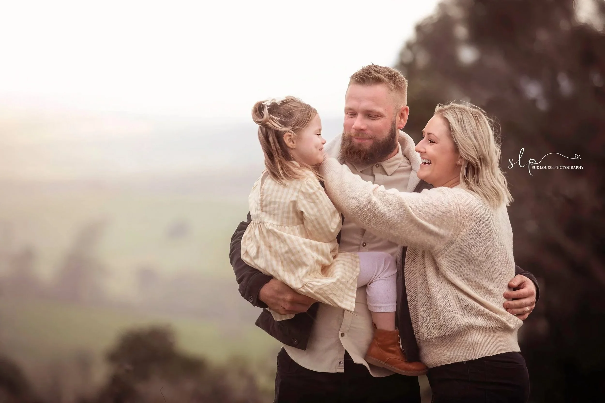 A family of three, a man, woman, and young girl, smiling and embracing outdoors during daytime, with trees and a cloudy sky in the background.