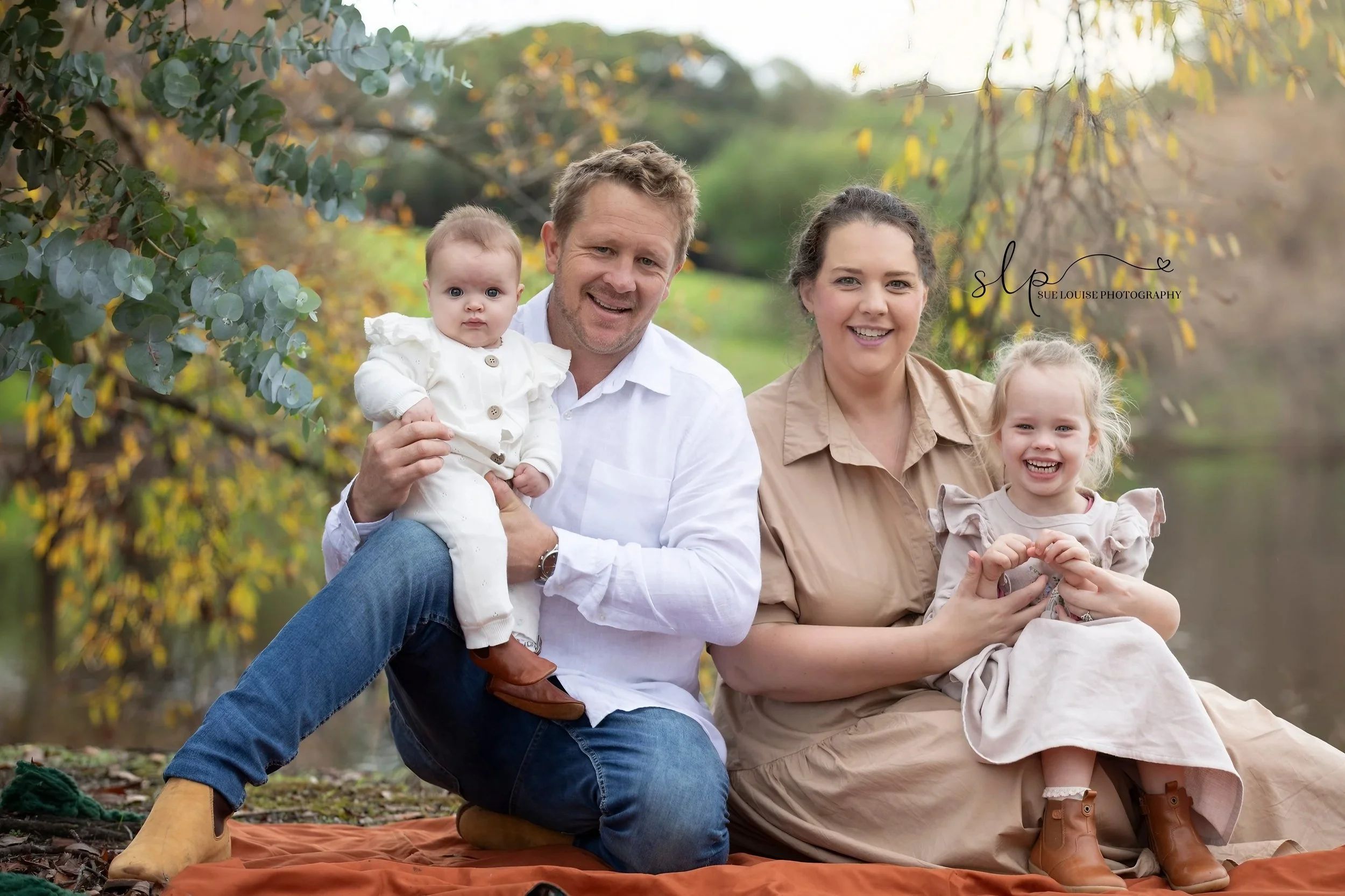 A family of four sitting outdoors by a river, smiling at the camera, with fall foliage in the background.