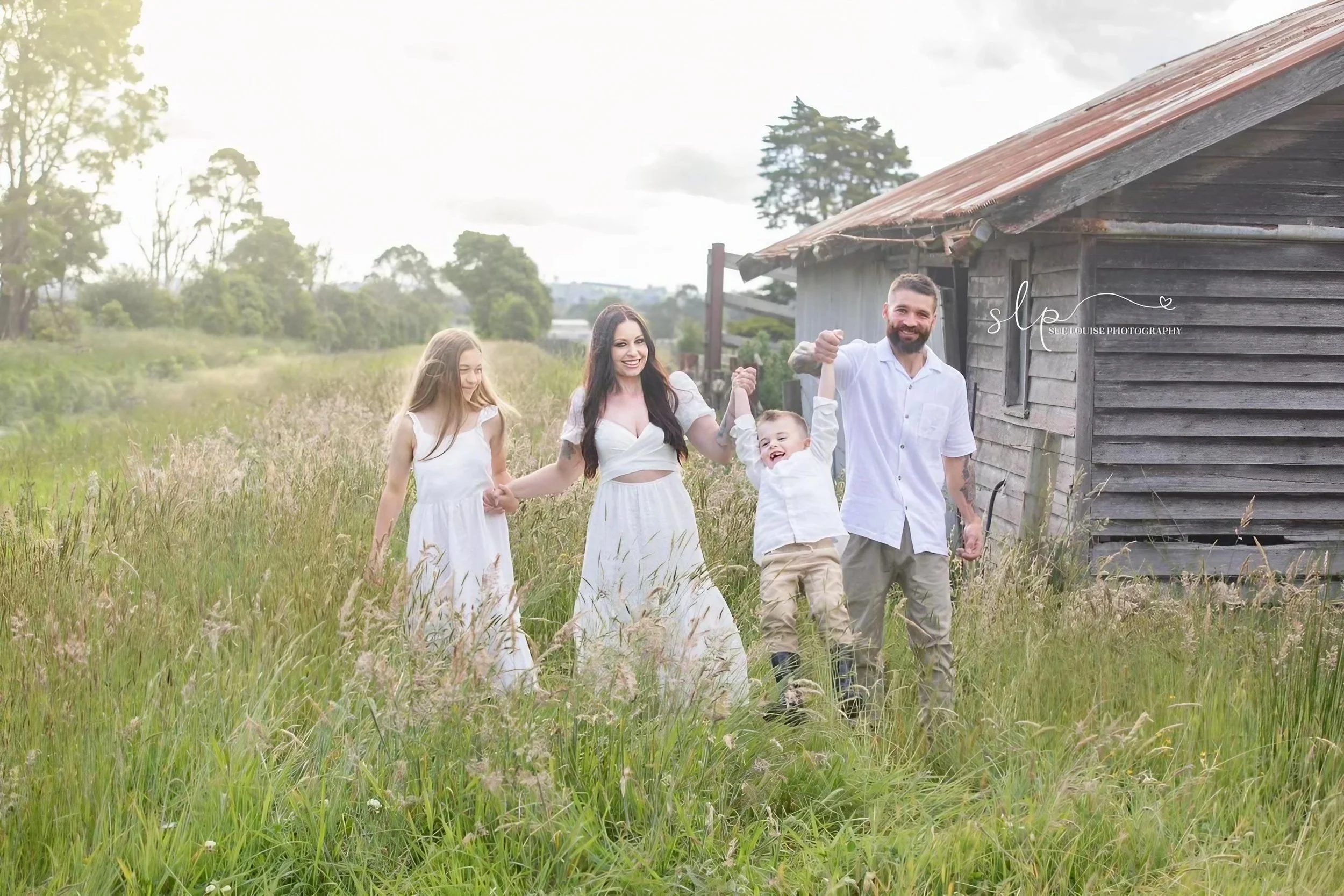 Family of four holding hands and smiling in a grassy field near an old wooden barn, wearing white clothes.