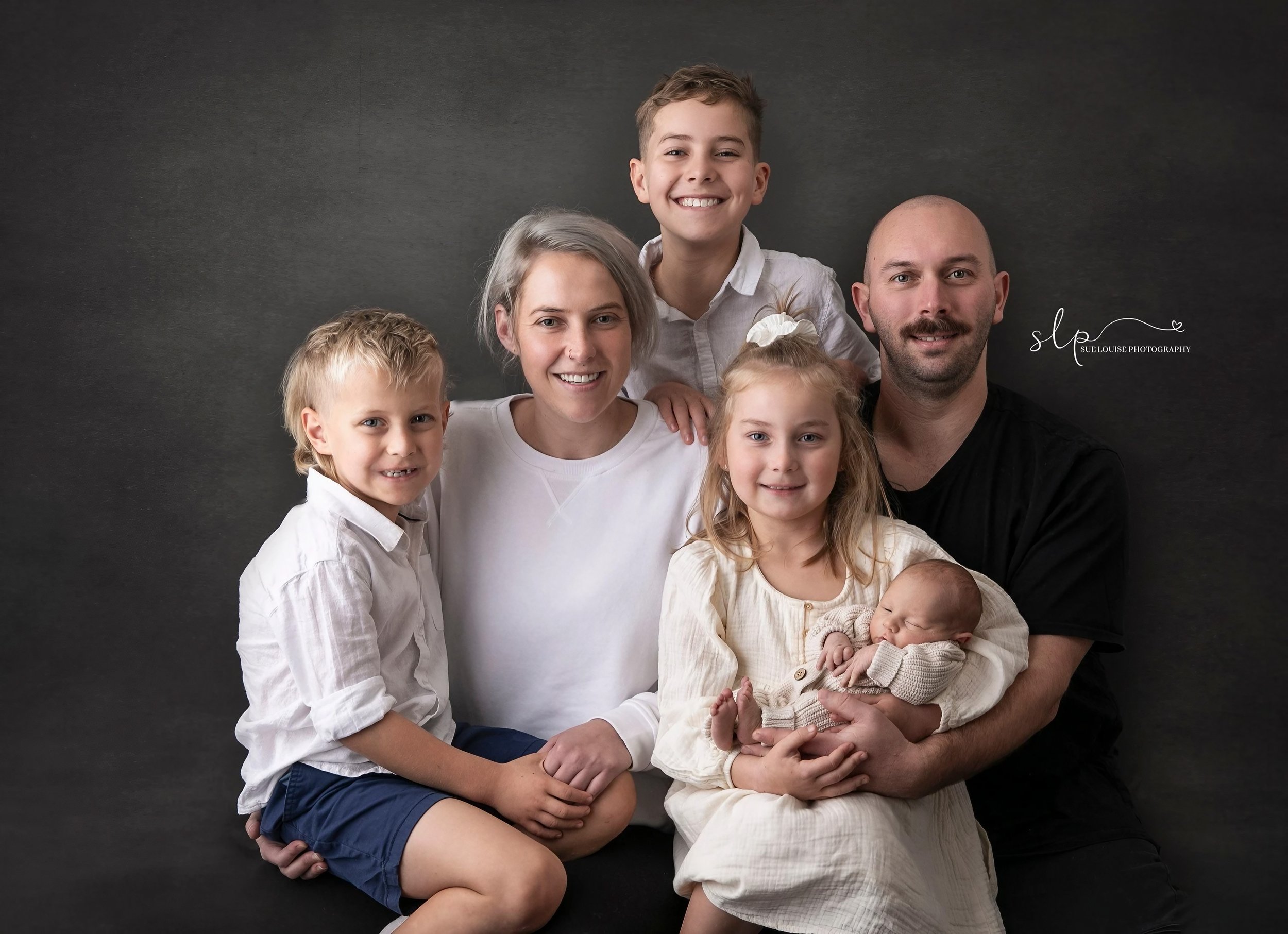 Family of six, with two children, a baby, and two adults, posing together in front of a dark background, smiling for a portrait photo.