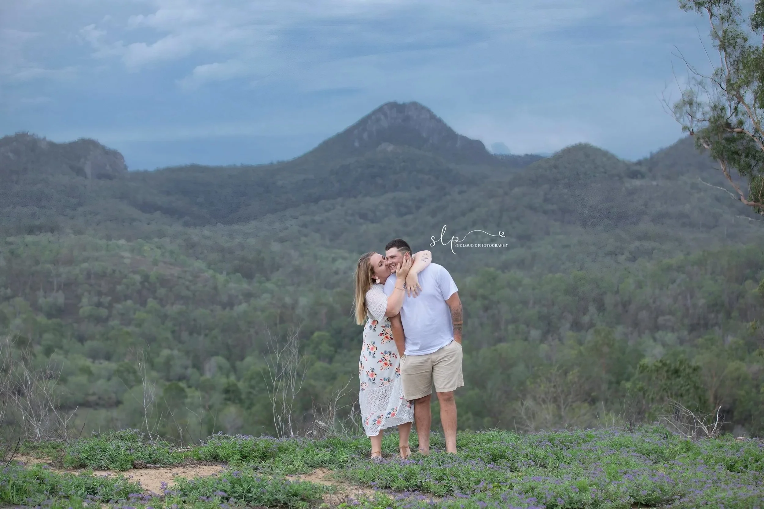 A couple hugs and kisses in a scenic outdoor setting with green hills and mountains in the background, under a partly cloudy sky.
