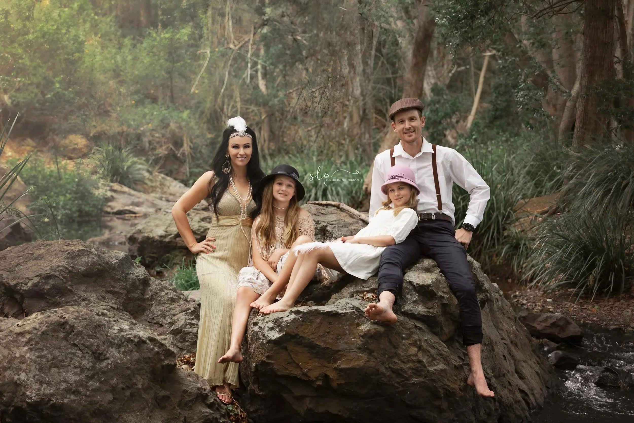 A family of five posing on rocks beside a creek in a wooded area, with the mother wearing a vintage dress and accessories, the father dressed in vintage-inspired white shirt and suspenders, and three young girls in vintage-style dresses and hats.