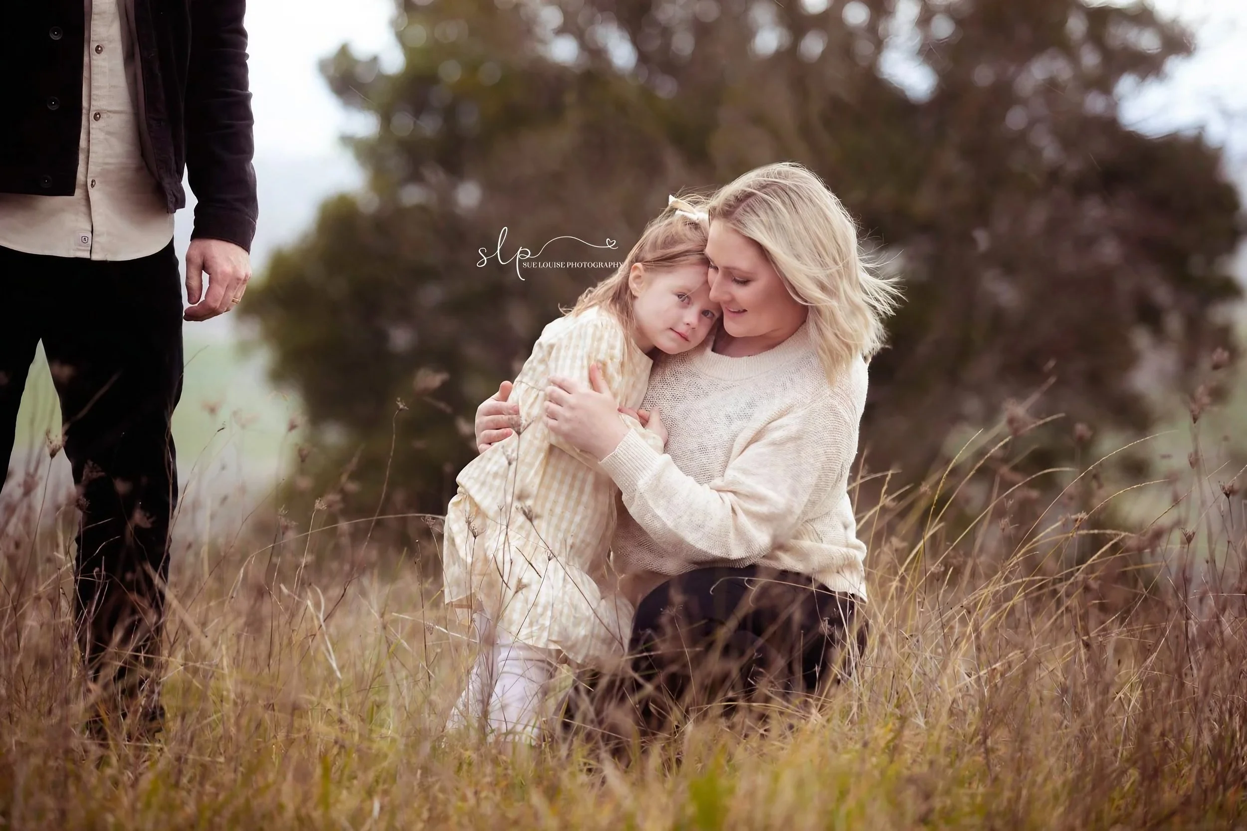 A woman with blonde hair, wearing a beige sweater, kneeling in a grassy field, hugging a young girl with light brown hair, wearing a yellow plaid dress. An adult male is standing nearby, partially visible on the left side of the image. There are tree