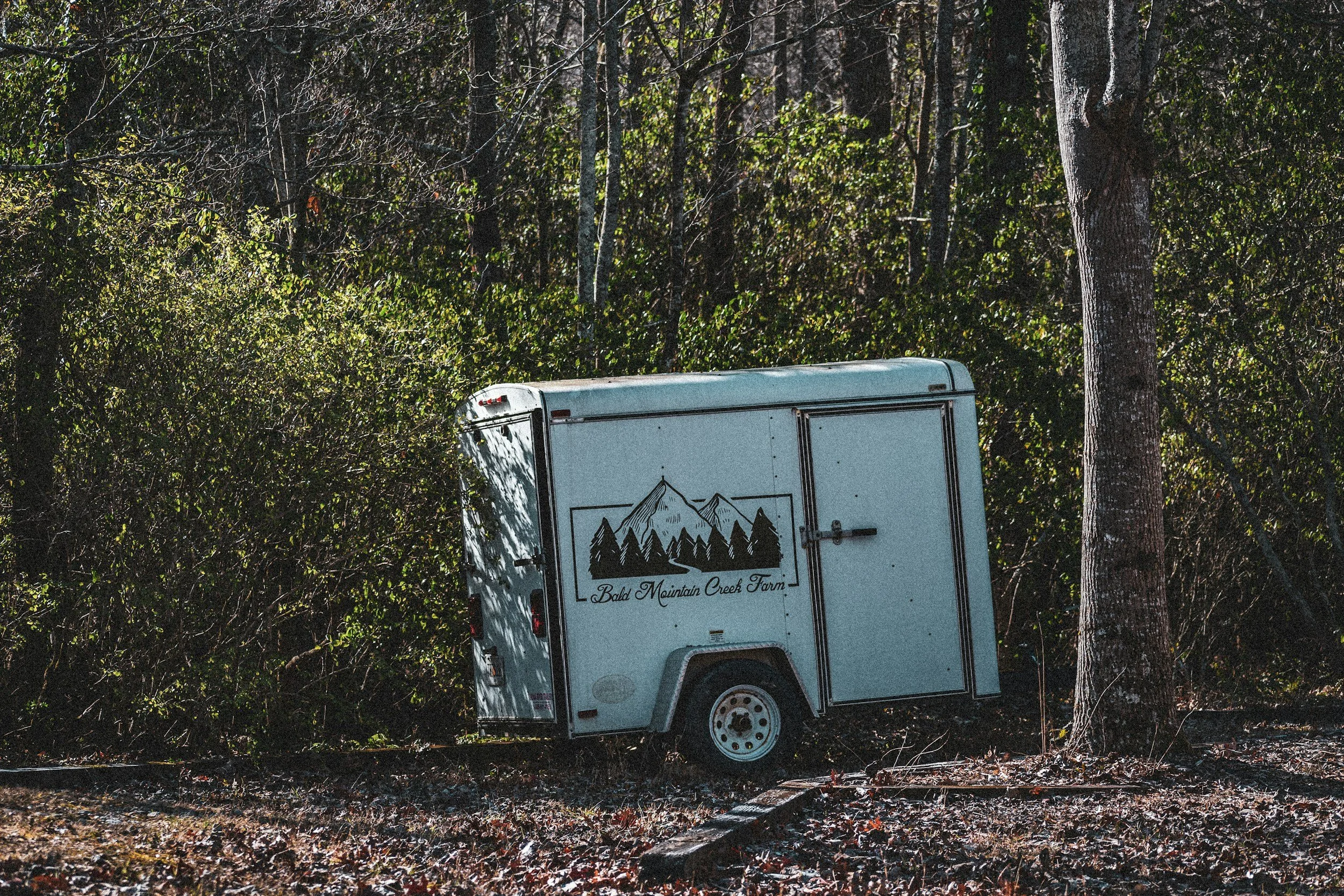 A white trailer with mountain and pine tree graphics and the text 'Bald Mountain Creek Farm' parked on grass next to a tree in a wooded area.
