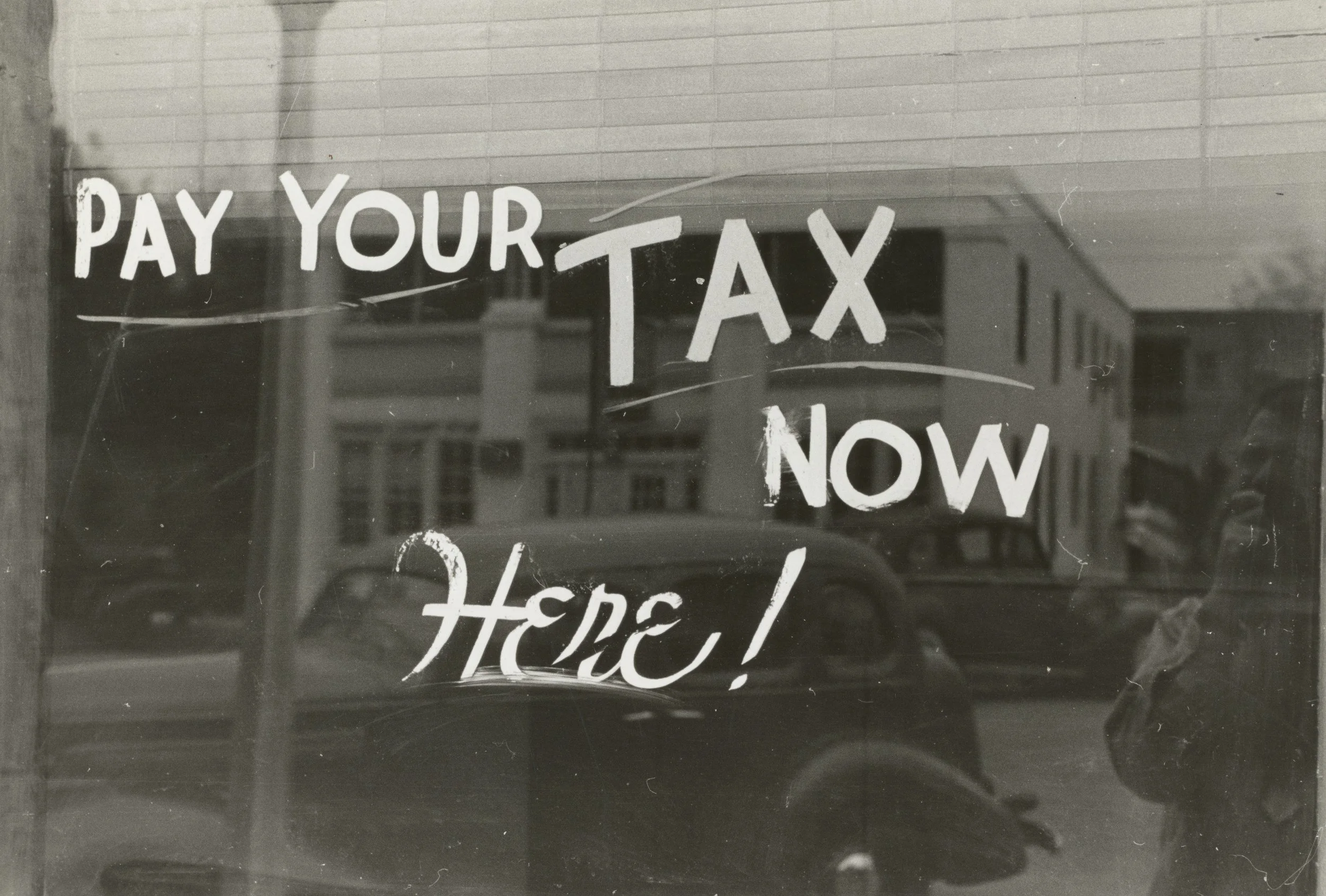 A sign on a window that says "Pay Your Tax Now Here!" in large, hand-painted letters.