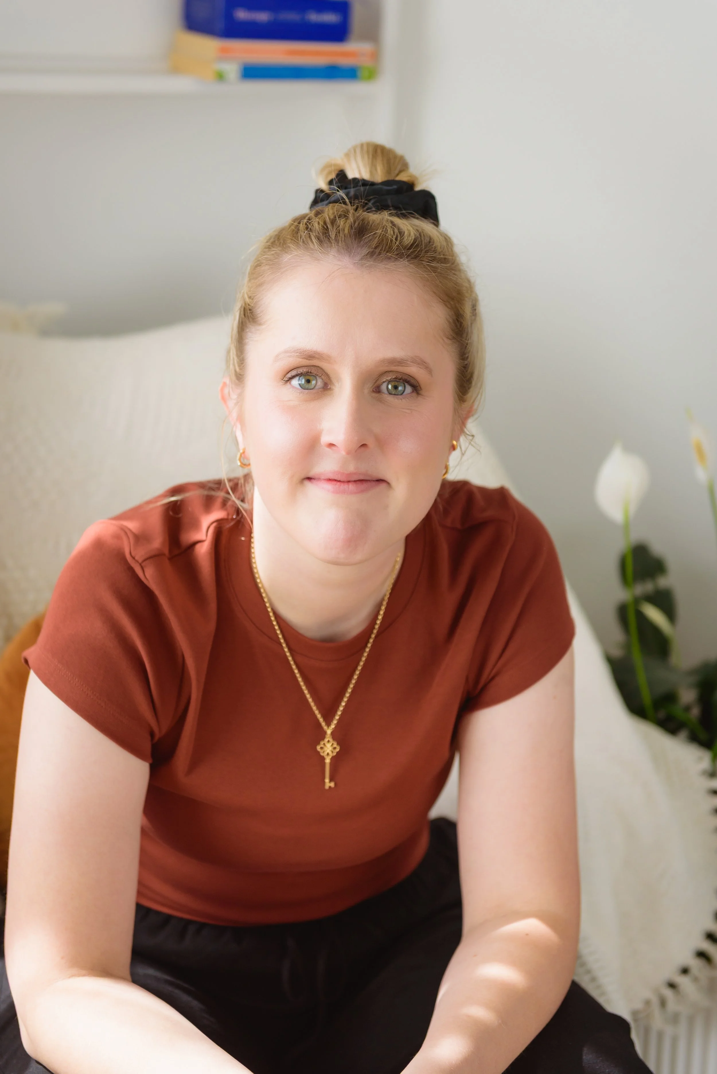 A woman with blonde hair tied in a bun, wearing a brown T-shirt and gold jewelry, sitting on a sofa in a bright room.