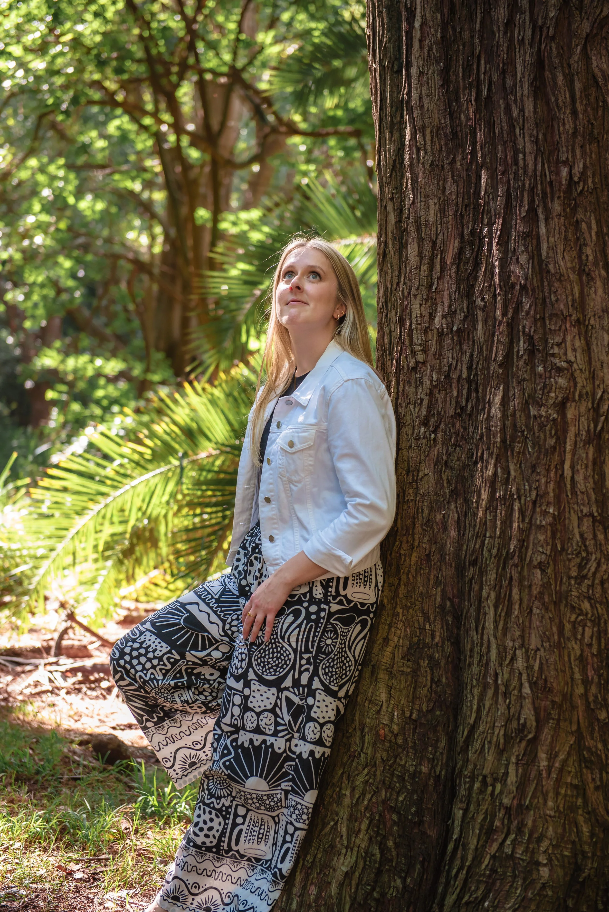 A young woman with long blonde hair, dressed in a white jacket and patterned black-and-white pants, standing in a lush green forest leaning against a large tree trunk, looking upwards.