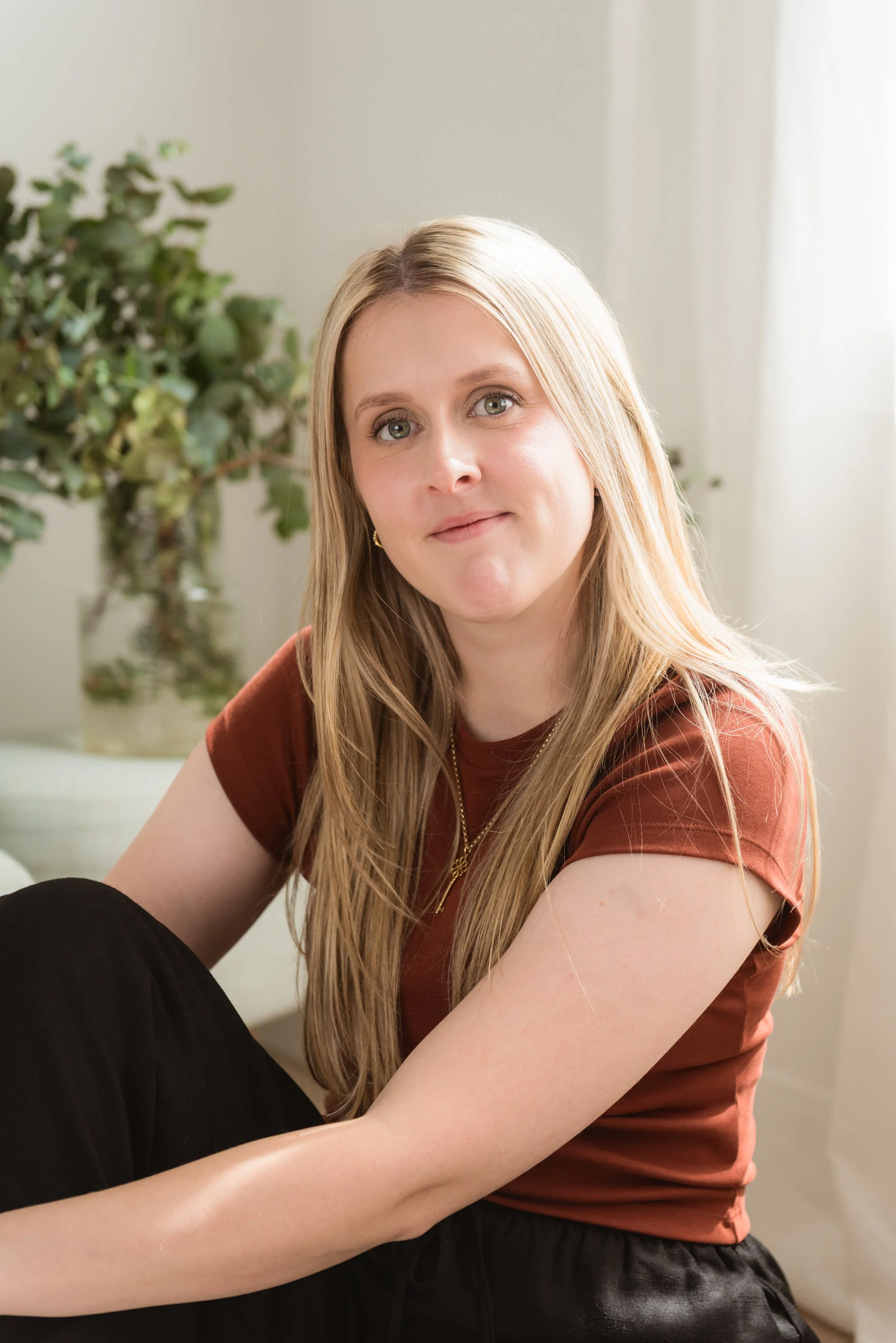 A young woman with long blonde hair, wearing a rust-colored t-shirt and black pants, sitting indoors in front of a potted plant with a white curtain in the background.