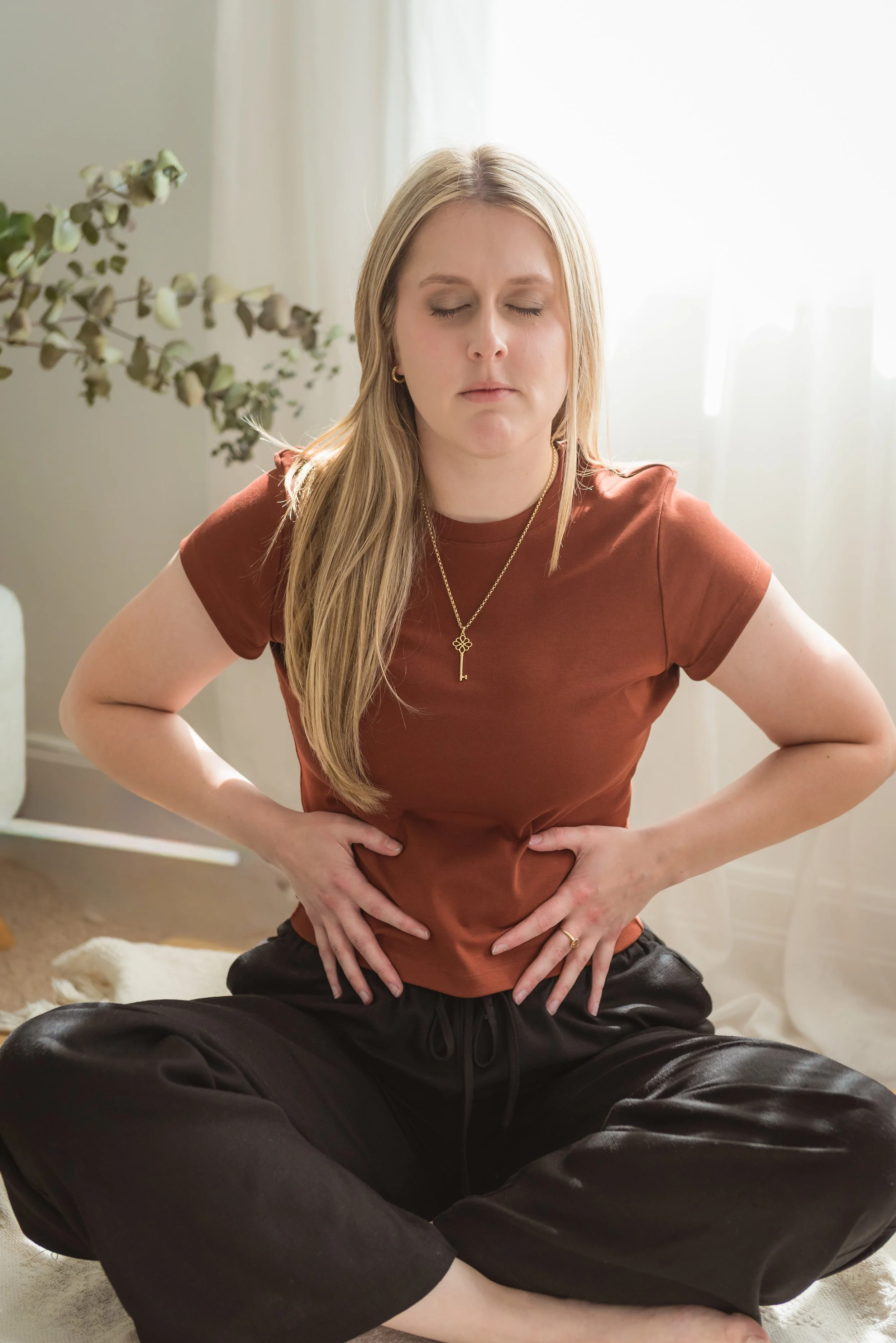 A woman sitting cross-legged on the floor with her eyes closed, holding her stomach with a pained expression.