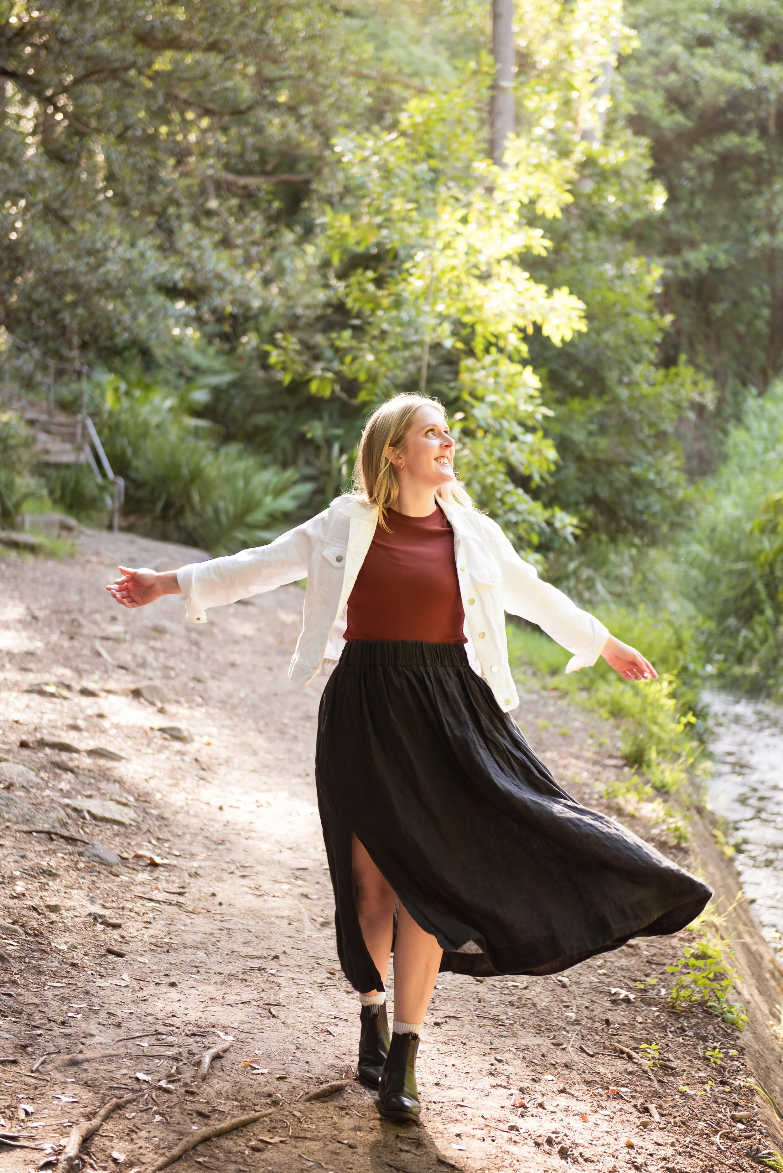 A woman dancing on a wooded trail with arms outstretched, wearing a white jacket, maroon top, black skirt with a slit, and black boots, near a river.
