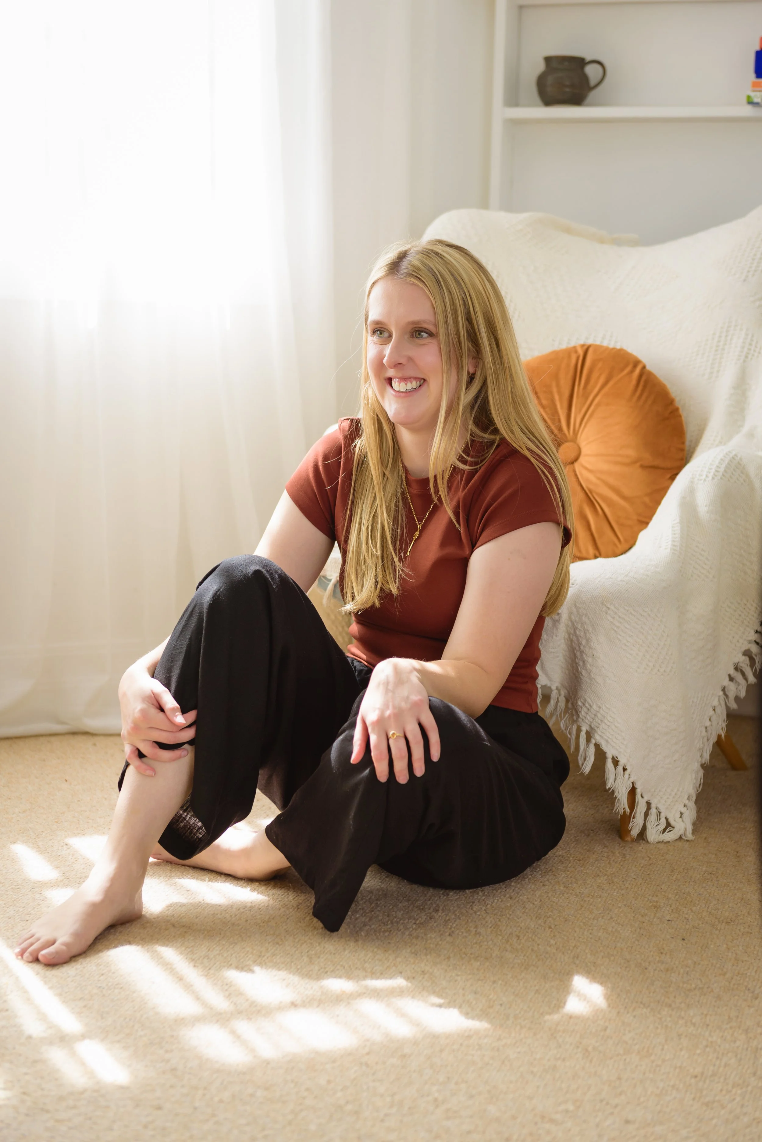 A woman sitting on a beige carpeted floor next to a white sofa with a round orange pillow, smiling and holding her leg, in a bright room with sunlight filtering through sheer curtains.