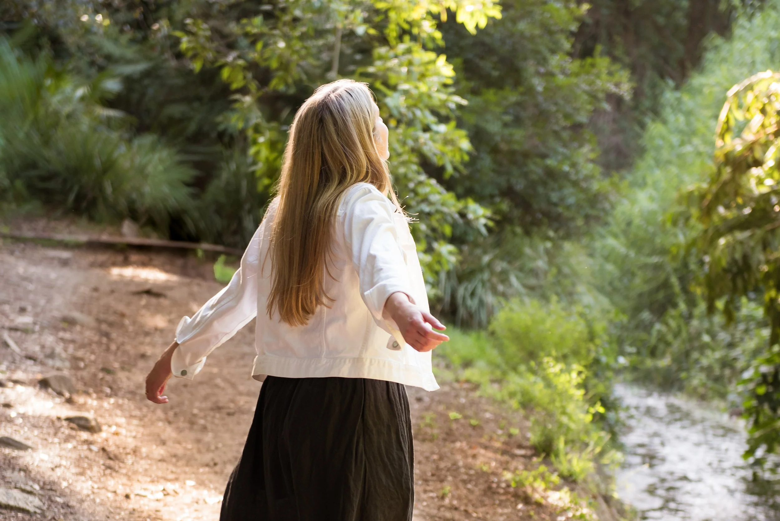 Woman with long blonde hair wearing a white jacket and black skirt walking on a forest trail beside a stream with lush green trees.
