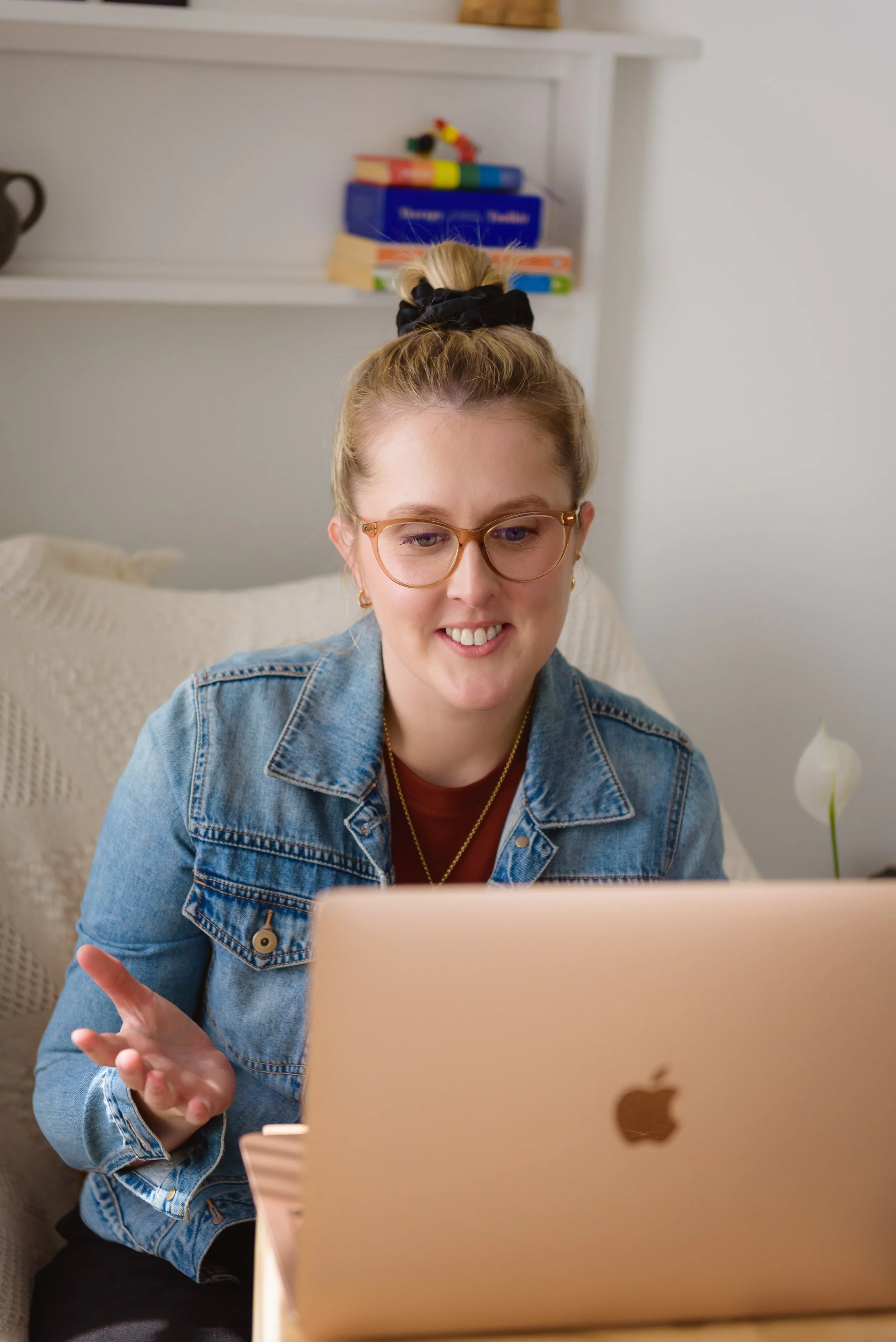 A young woman with glasses, wearing a denim jacket, smiling while looking at her laptop.