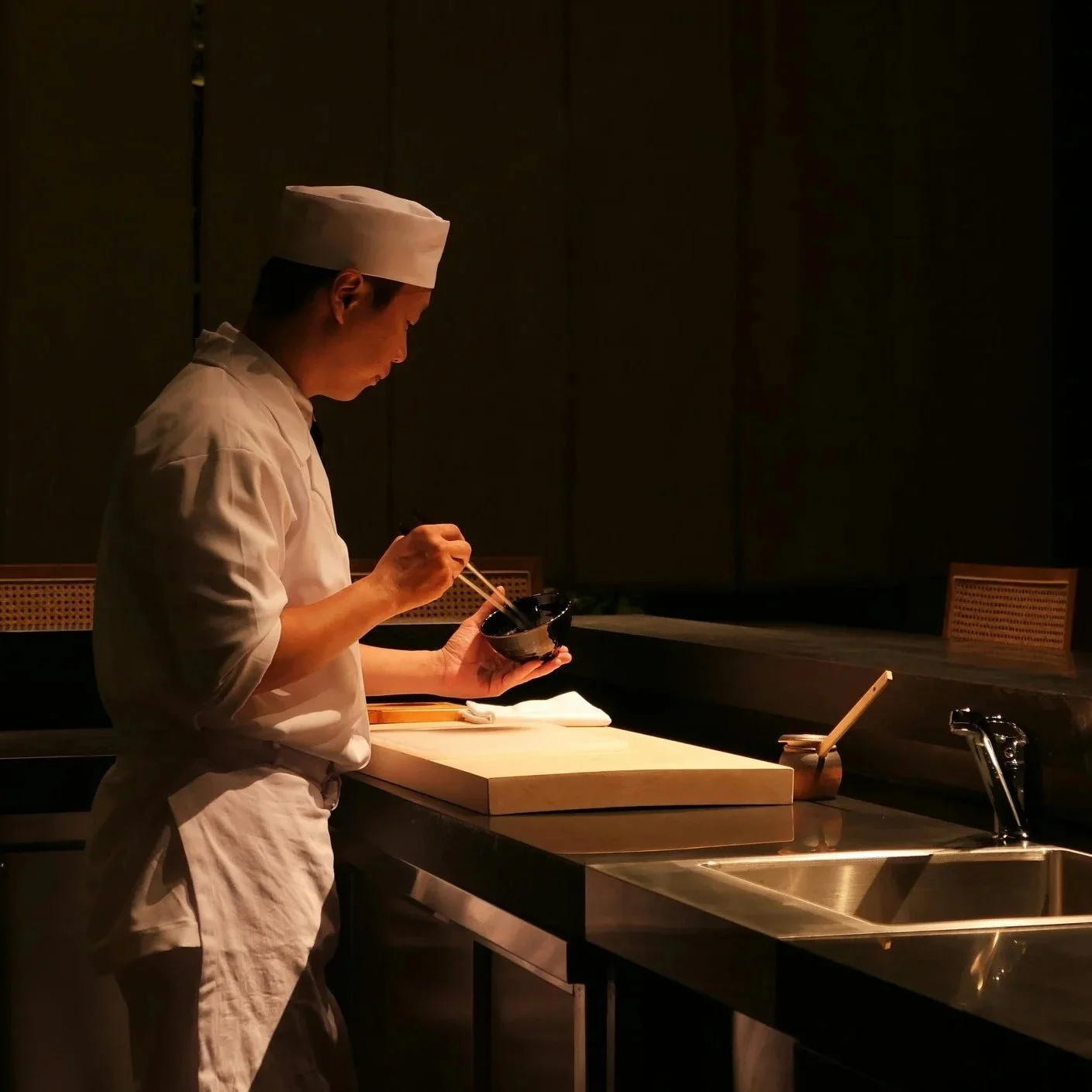 Chef holding chopsticks and standing beside a cutting board in a restaurant kitchen.